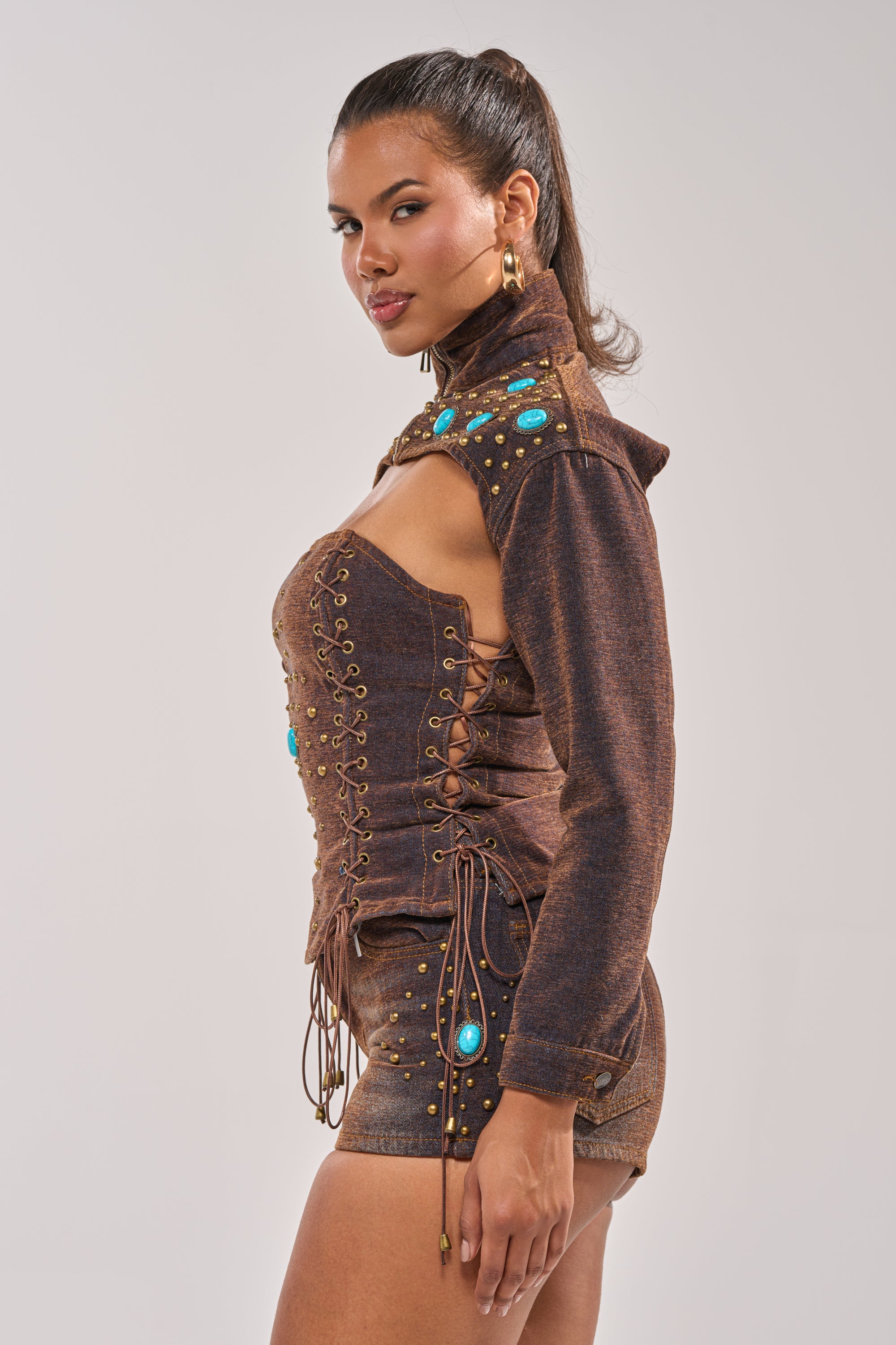 A woman poses in the INDIGO STUDDED DENIM BOLERO, a western-inspired, lace-up outfit with turquoise gemstones, gold studs, high collar, long sleeves, shorts, and her hair styled in a high ponytail. 3