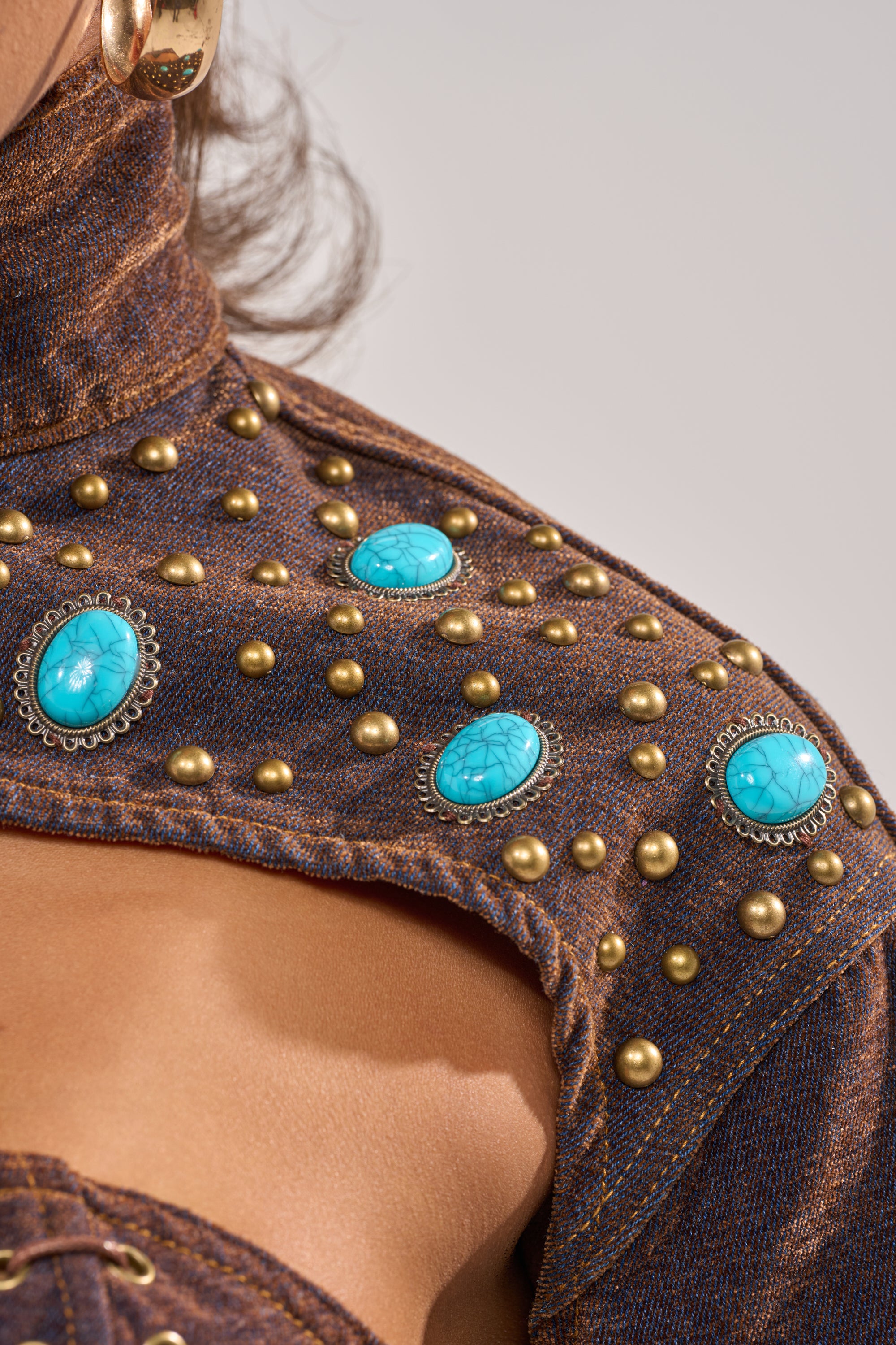 A close-up of a person wearing the INDIGO STUDDED DENIM BOLERO, featuring gold studs and oval turquoise gemstones on the shoulder, paired with a large gold hoop earring. 6