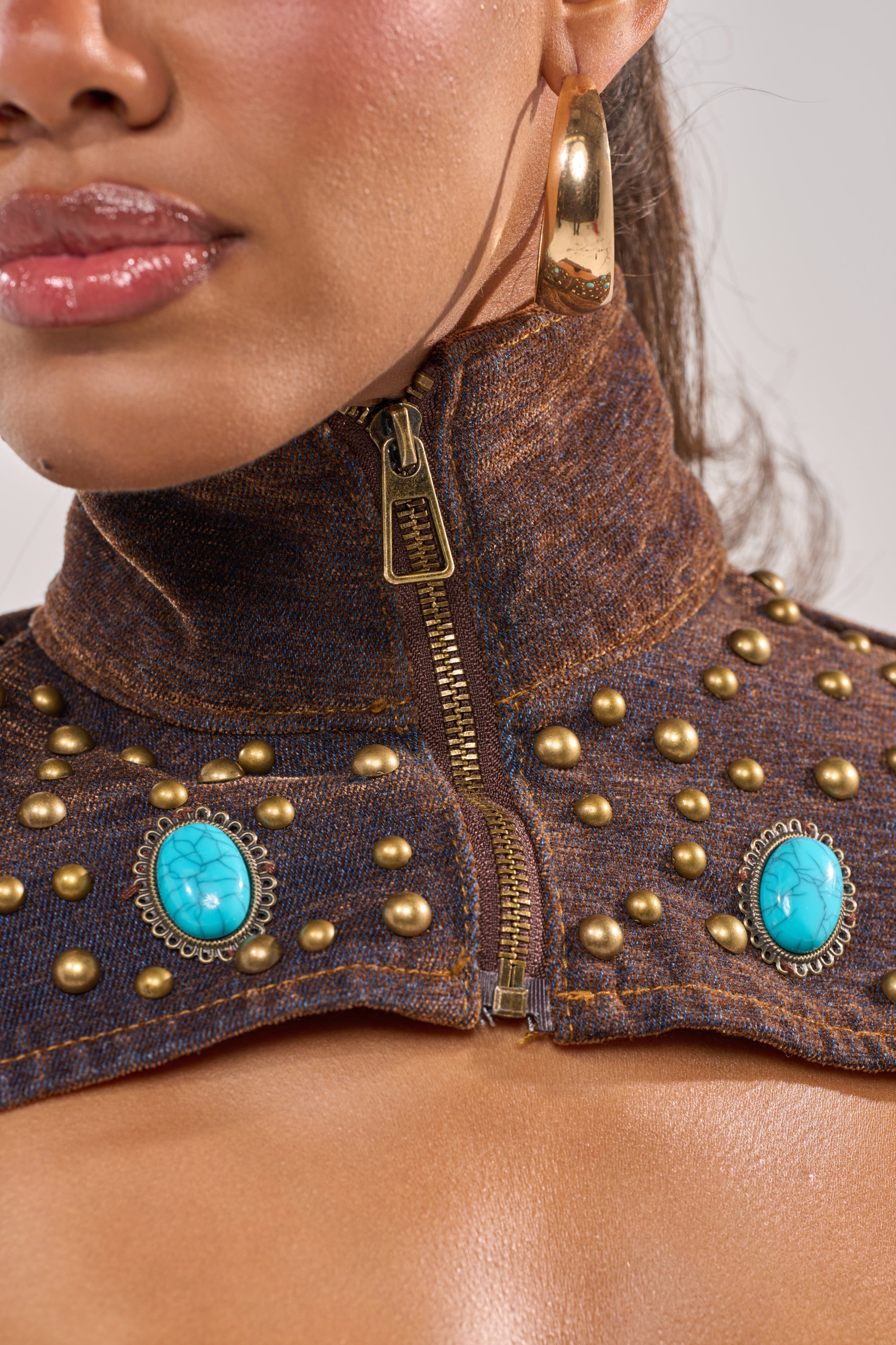 A close-up of a woman wearing the INDIGO STUDDED DENIM BOLERO, adorned with gold studs, turquoise brooches, a visible zipper, large gold hoop earrings, and glossy pink lipstick. 7