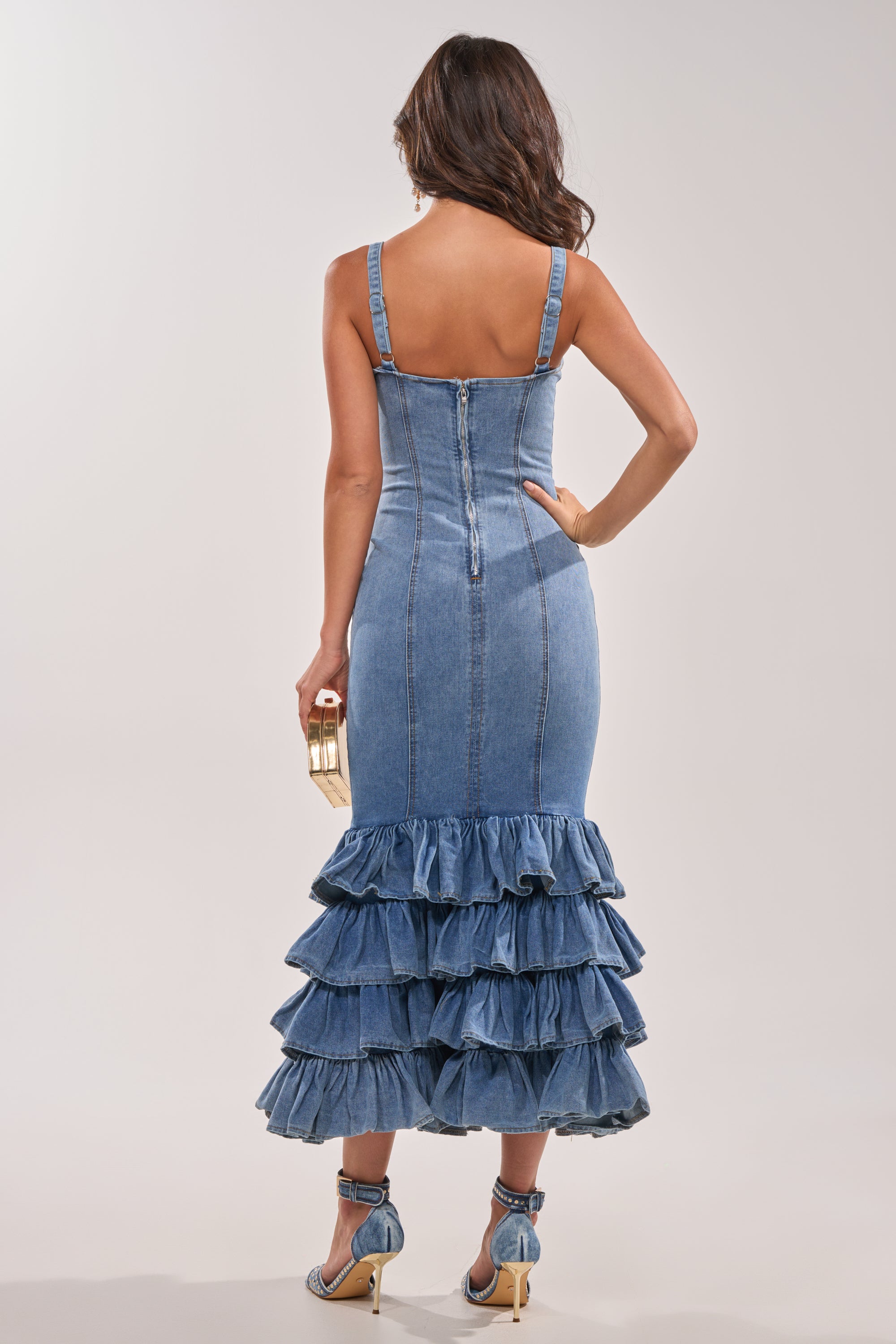 A woman faces away wearing the MAIN ONE DENIM RUFFLE MAXI DRESS with a tiered ruffle hem. She holds a gold clutch and wears strappy high-heeled sandals against a plain light-colored background. 3