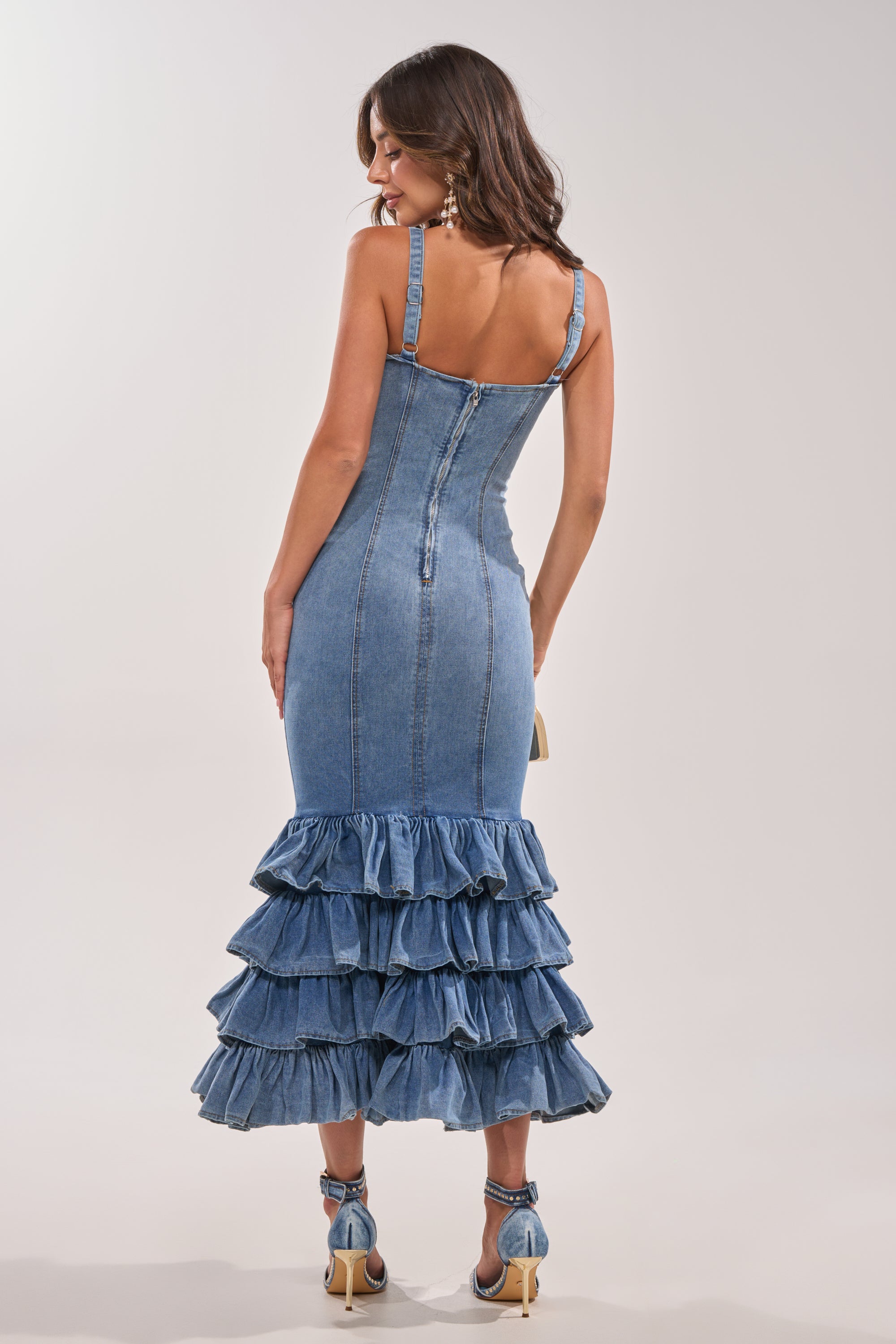 A woman faces away wearing the MAIN ONE DENIM RUFFLE MAXI DRESS featuring thin straps, a back zipper, and a tiered ruffle hem. She pairs it with heeled sandals and carries a small purse. 4