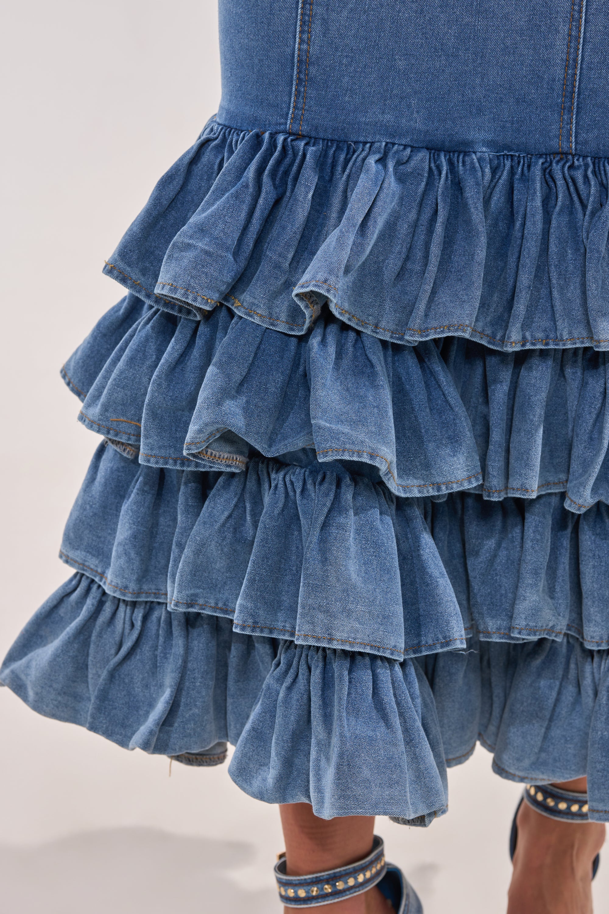 Close-up of the MAIN ONE DENIM RUFFLE MAXI DRESS, featuring tiered ruffled blue denim layers reminiscent of mermaid-inspired style. The hem is above the knee, paired with a sandal with denim straps partially visible below. 6