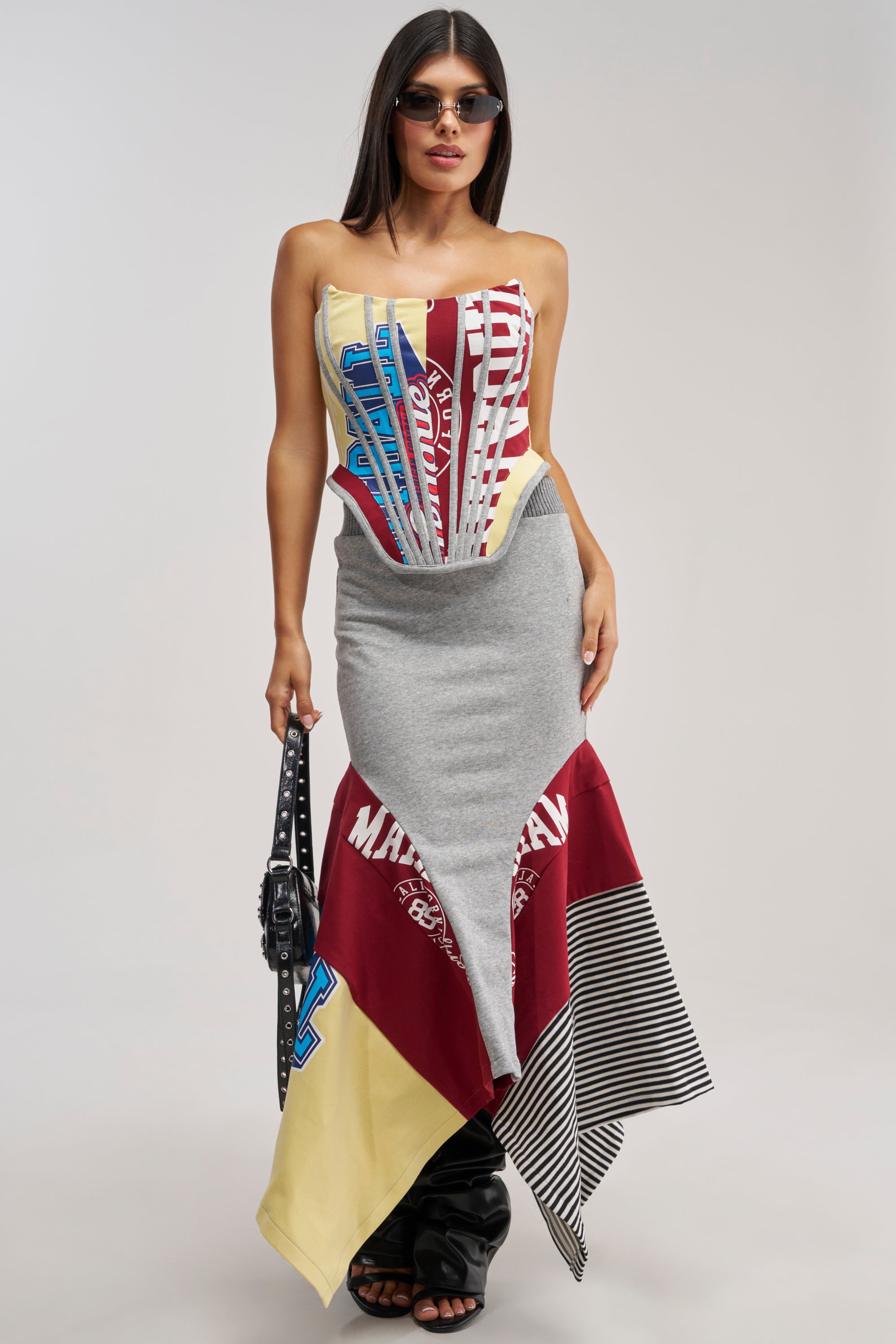 A woman wearing sunglasses pairs the GOING FOR IT ALL MIDI SKIRT—a gray and red varsity mermaid silhouette—with a strapless patchwork logo dress, black studded handbag, and black shoes, effortlessly showcasing elevated streetwear style against a neutral background.