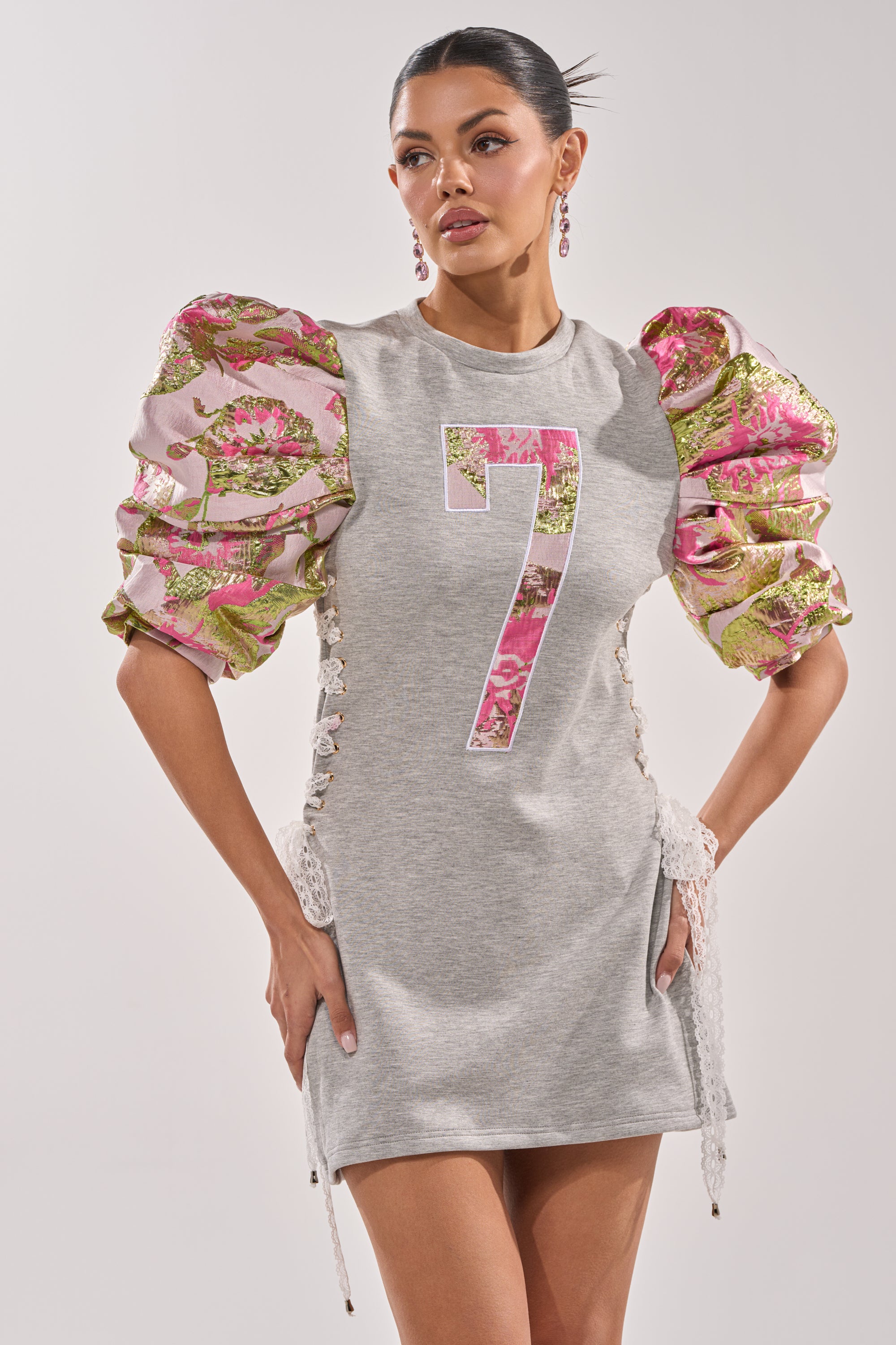A woman models the GAME CHANGER BROCADE LACE UP MINI DRESS, a short grey dress featuring a bold pink floral number 7 and puffed floral-pattern sleeves, standing with hands on hips and gazing to the side. 0