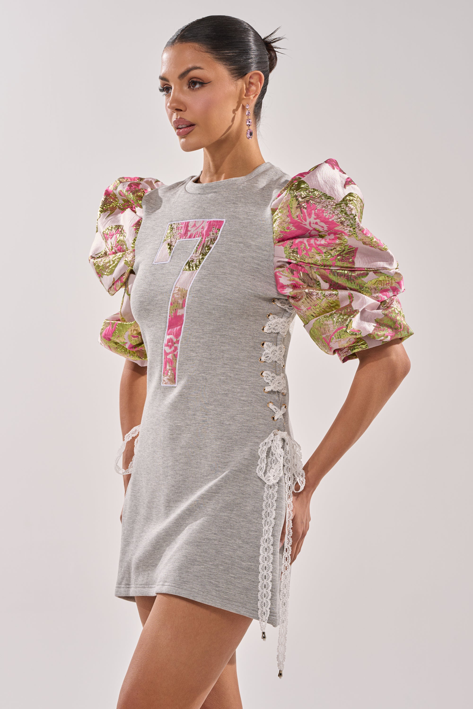 A woman models the GAME CHANGER BROCADE LACE UP MINI DRESS in heather gray with brocade puff sleeves, lace-up sides, and a bold pink number 7, posing against a plain light background. 3