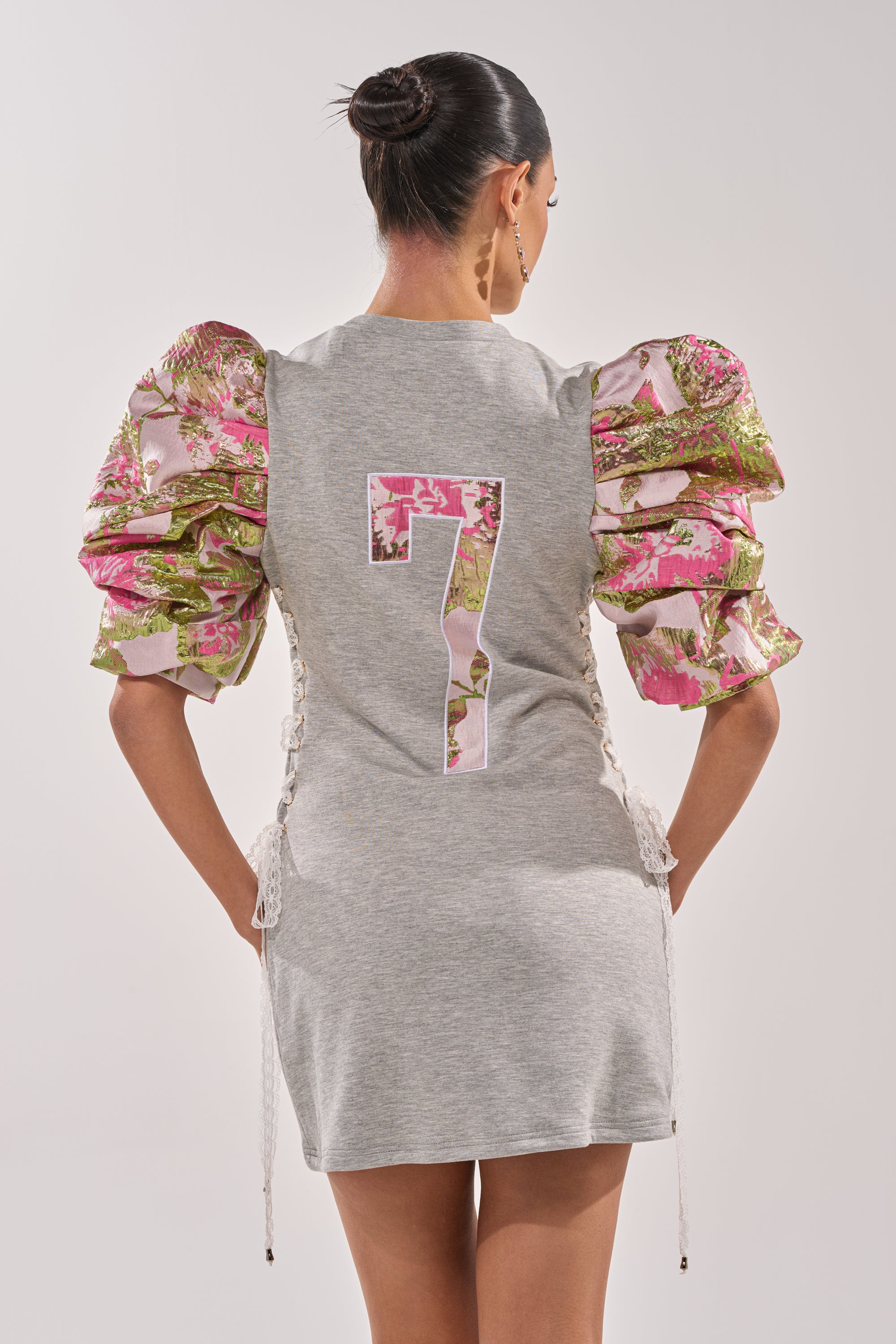 A woman faces away wearing the GAME CHANGER BROCADE LACE UP MINI DRESS in heather gray with pink and green brocade puff sleeves and a floral-outlined number 7 on the back. Her hair is in a neat bun for game-day glam. 4