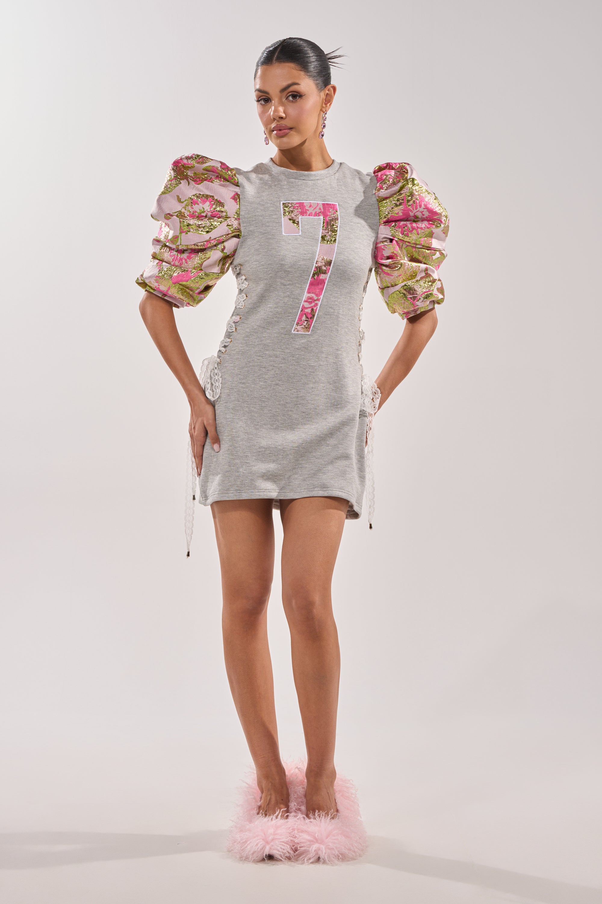 A woman wears the GAME CHANGER BROCADE LACE UP MINI DRESS in heather gray with a bold "7" on the front, floral puffed sleeves with brocade details, and pink fluffy slippers, standing against a plain light background. 5