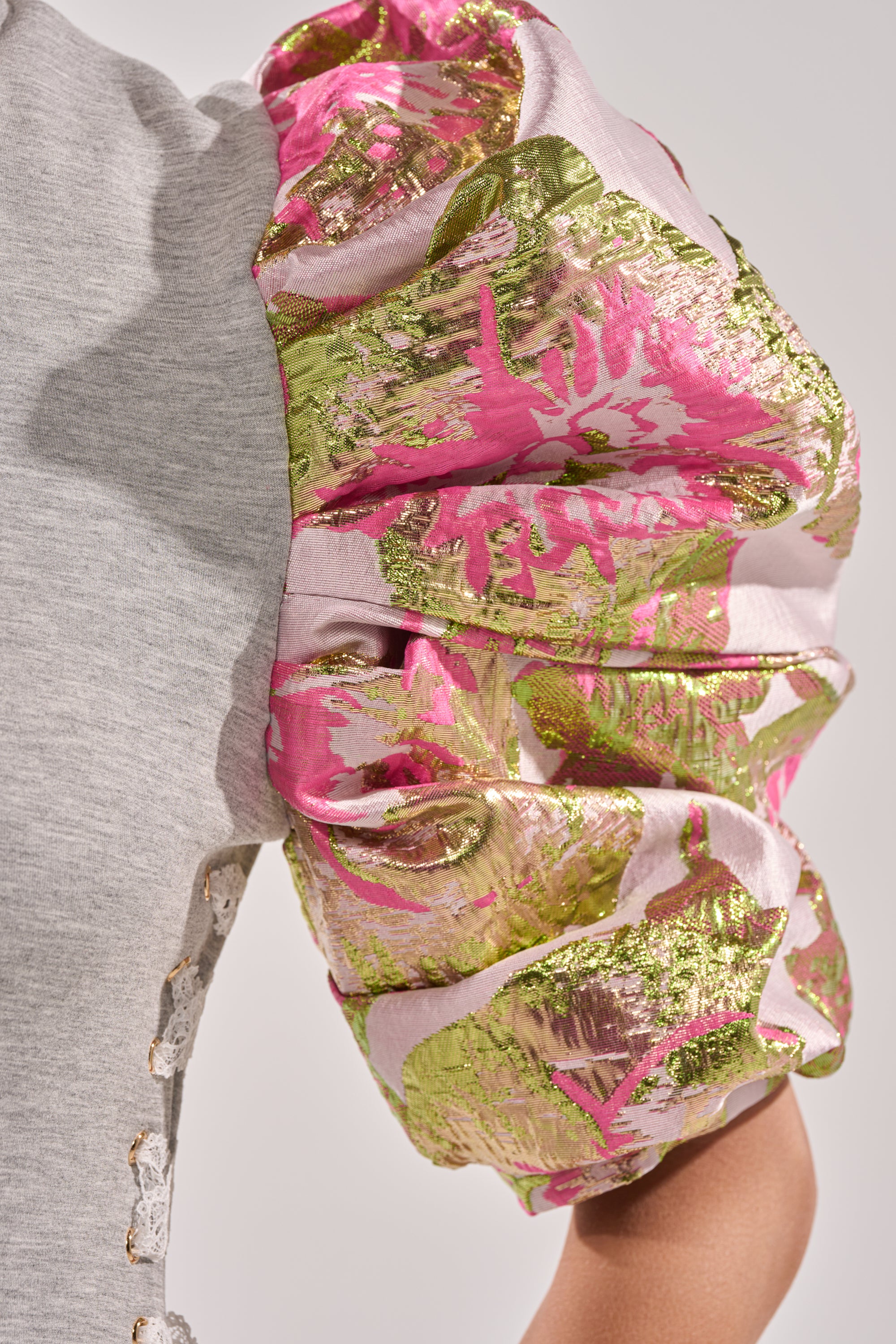 Close-up of the GAME CHANGER BROCADE LACE UP MINI DRESS with shiny metallic puff sleeves, pink, green, and gold floral brocade details, light heather gray body fabric, and decorative buttons along the seam. 7