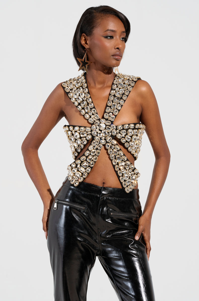 A person confidently showcases a striking fashion look featuring the RHINESTONE ENCRUSTED DRAMATIC TOP with a plunging neckline, complemented by shiny black leather pants. They accessorize with large star-shaped earrings, standing against a plain background.