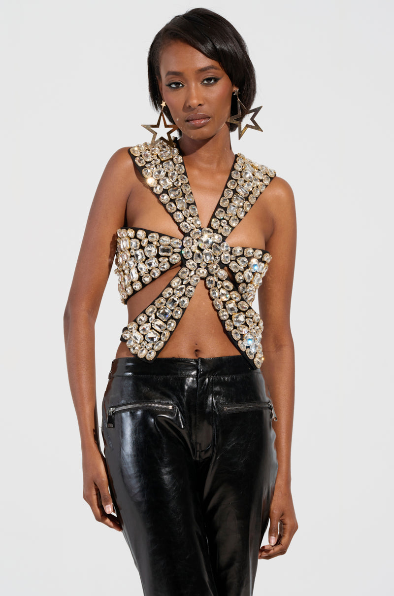 A person stands confidently in a unique outfit with large star earrings, wearing the Rhinestone Encrusted Dramatic Top that showcases sexy rib cage cut-outs and gleaming stones in a cross pattern, paired with sleek black pants.
