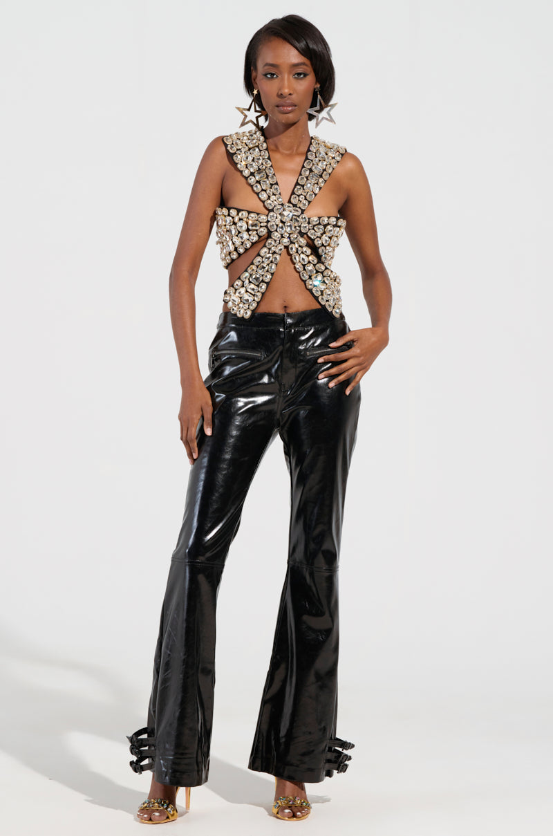 A woman confidently showcases the RHINESTONE ENCRUSTED DRAMATIC TOP, which features a plunging neckline and sexy rib cage cut-outs. She pairs this unique piece with shiny black flared pants and large star-shaped earrings, completing her look with high heels against a plain white background.