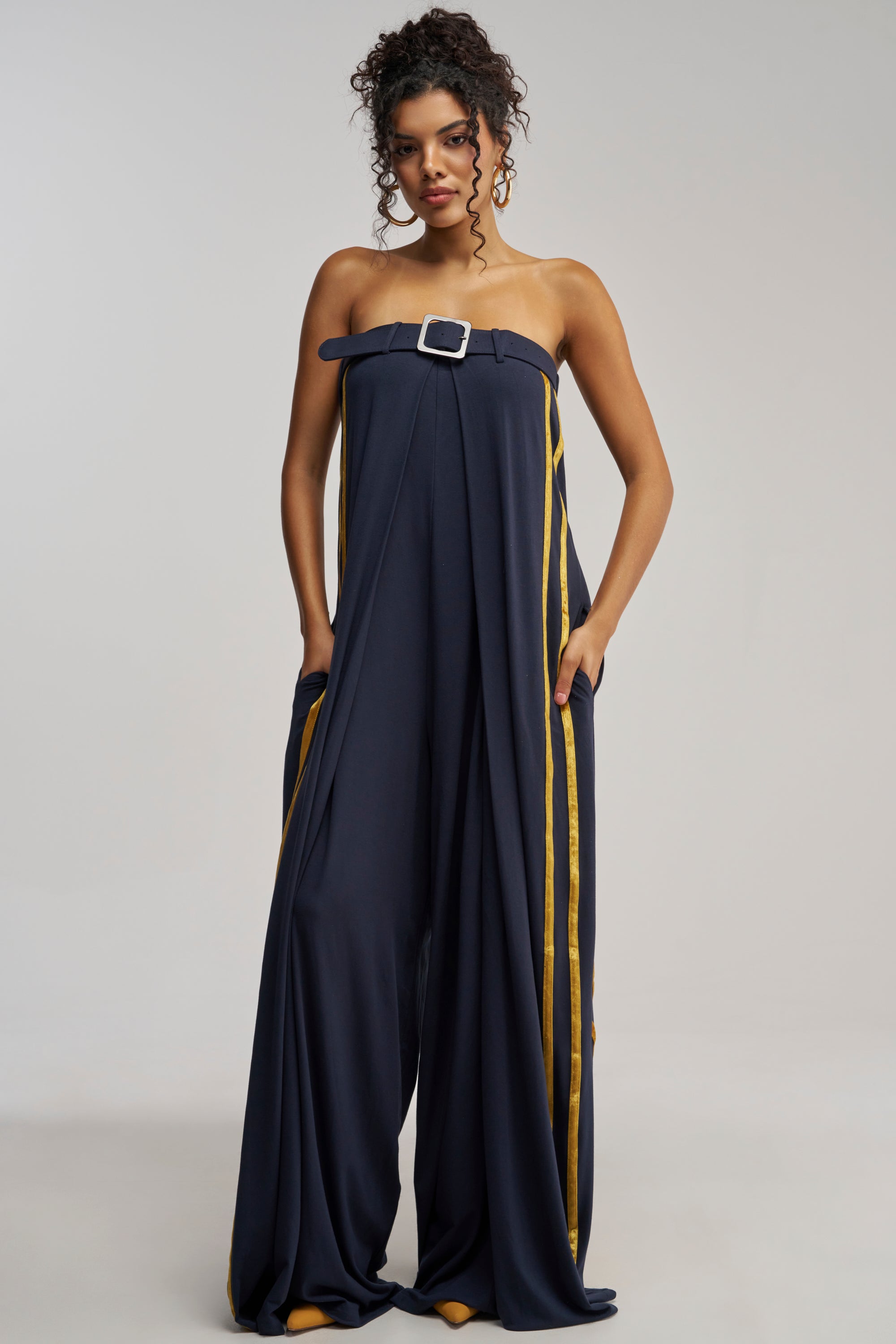 Wearing the RUN THE SHOW JUMPSUIT IN NAVY—featuring a strapless wide-leg silhouette, bold yellow side stripes, a belted waist with a silver buckle, and paired with yellow shoes—a woman stands hands in pockets against a plain backdrop for an effortlessly chic summer date night look.