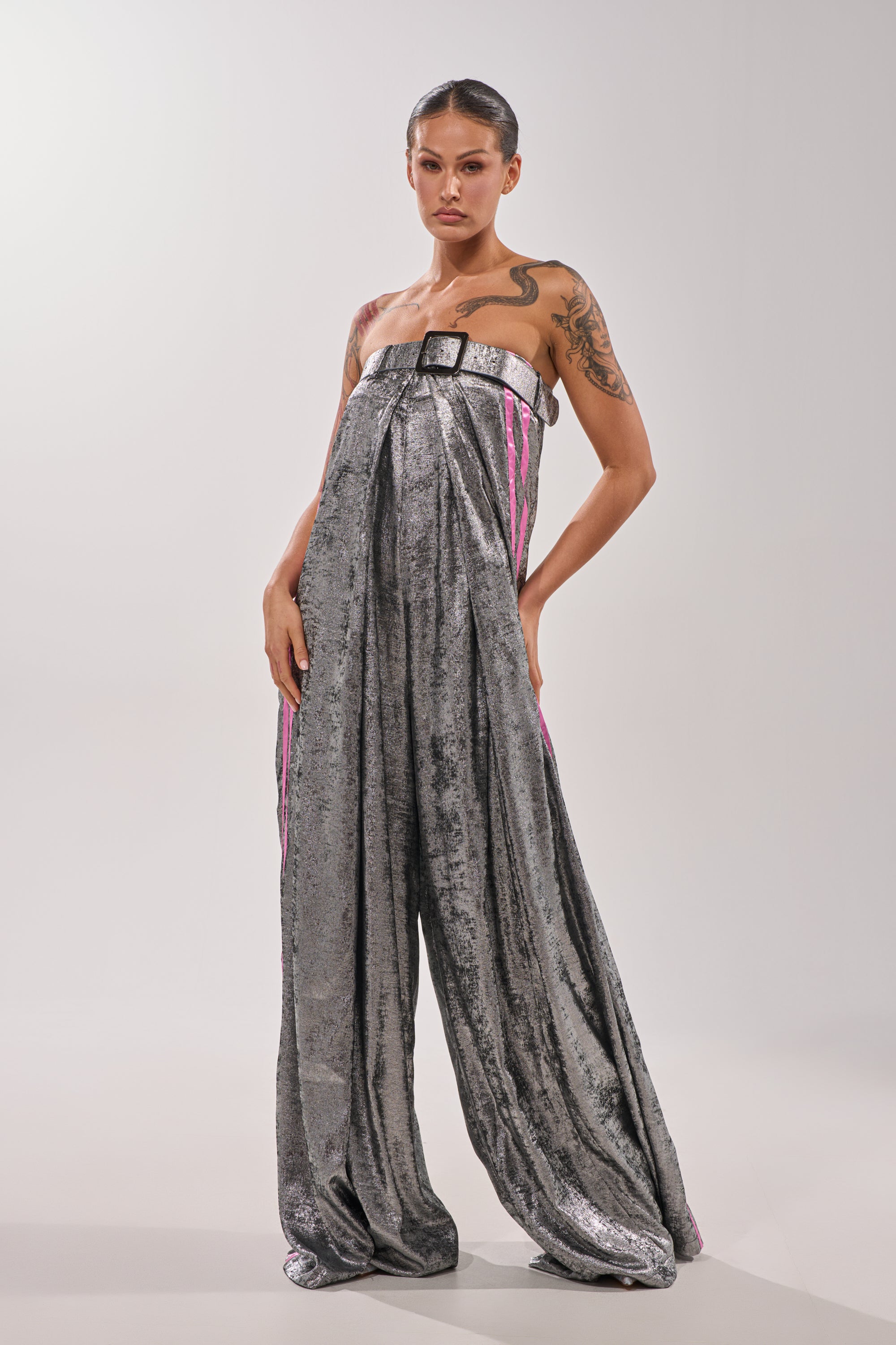 A tattooed individual models the RUN THE SHOW METALLIC JUMPSUIT—strapless, wide-legged, with silver sequins, black buckle detail, and pink side accents—standing confidently against a plain light background.