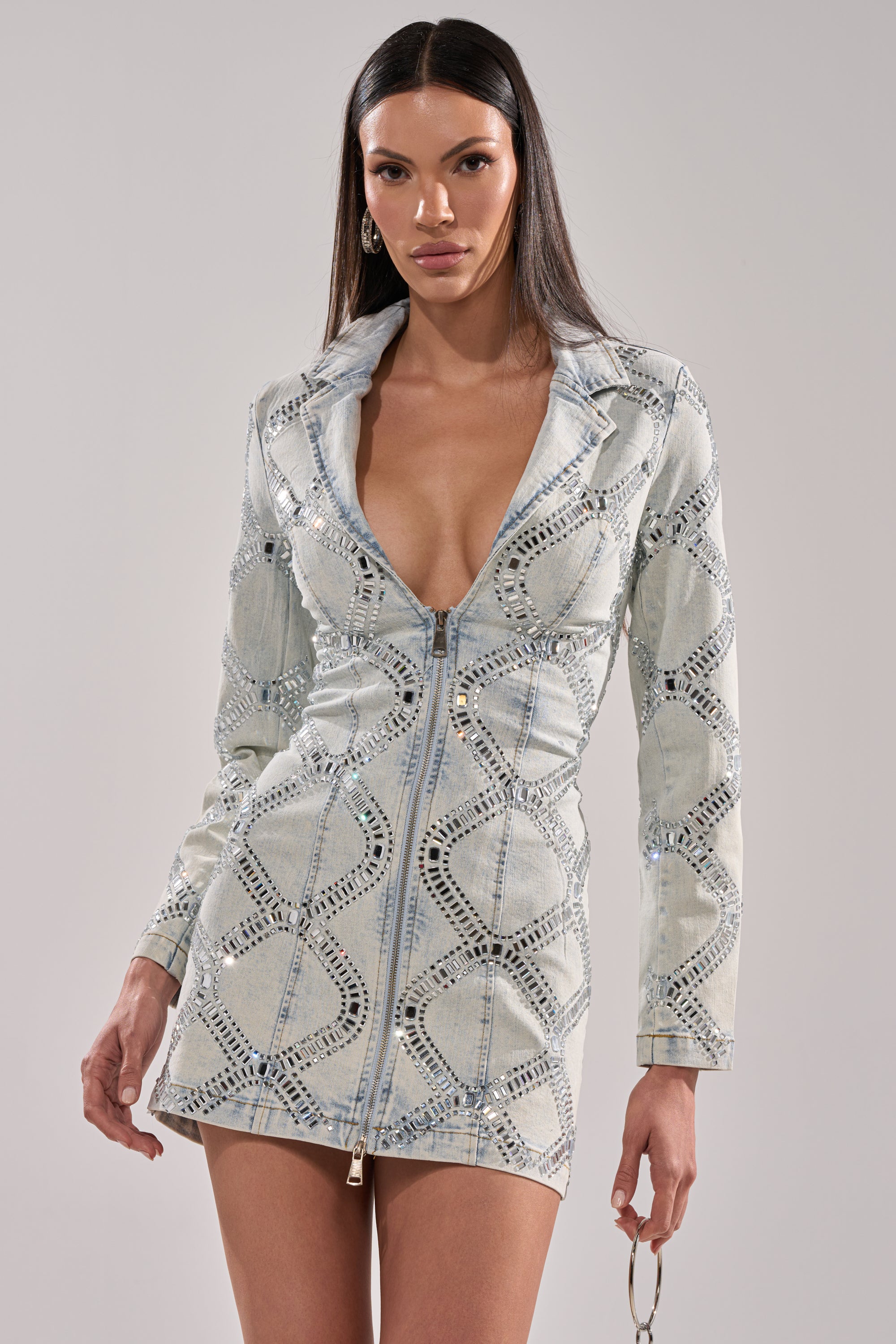 A woman poses against a plain background wearing the LOOK AT ME RHINESTONE BLAZER DRESS IN LIGHT BLUE DENIM, featuring long sleeves, a plunging neckline, zipper front, and geometric rhinestone pattern. She holds round sunglasses.