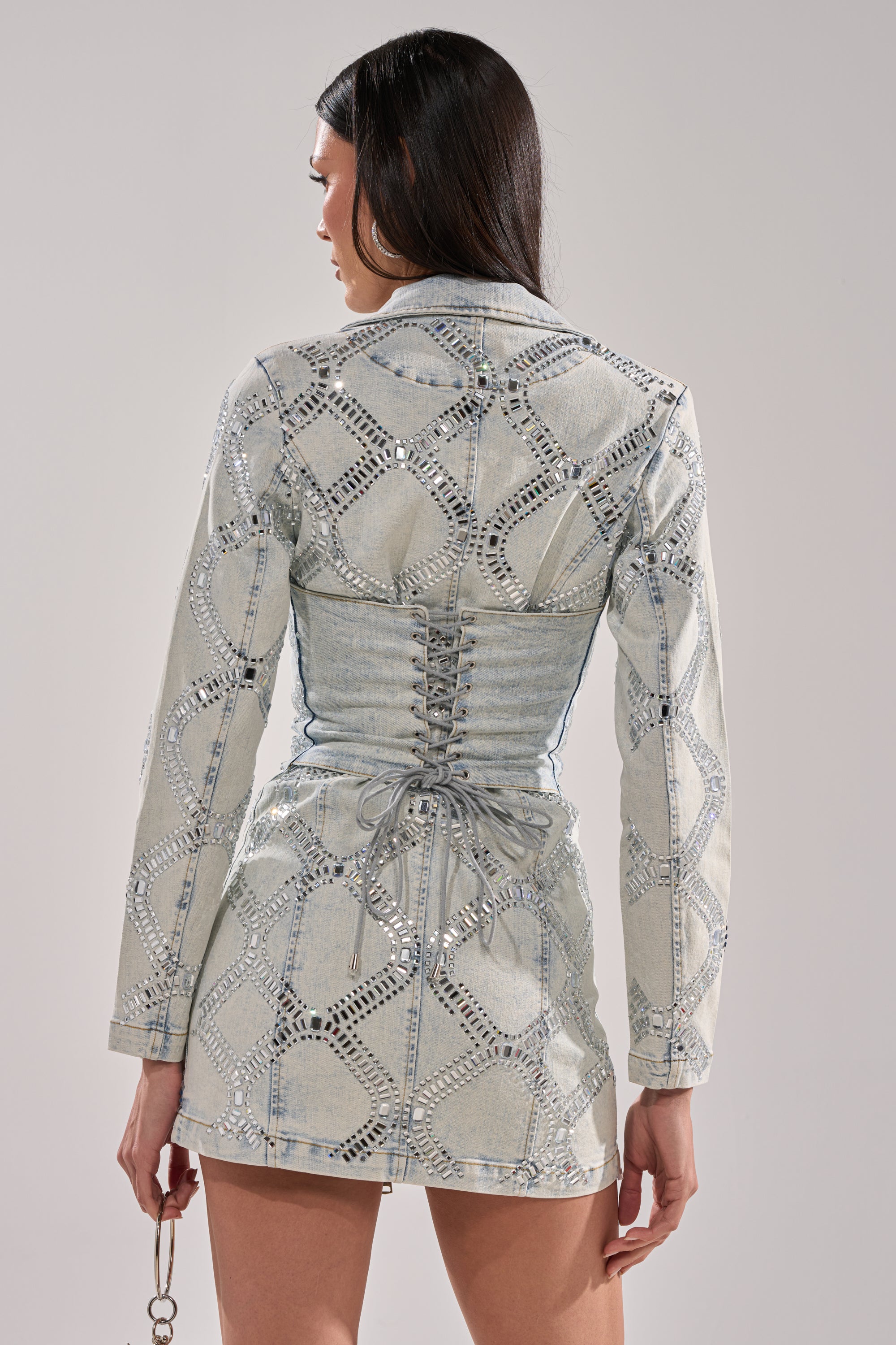 A woman faces away wearing the LOOK AT ME RHINESTONE BLAZER DRESS IN LIGHT BLUE DENIM, featuring a fitted mini silhouette, long sleeves, silver studded geometric patterns, and a lace-up corset back detail. She holds a small item in her left hand.