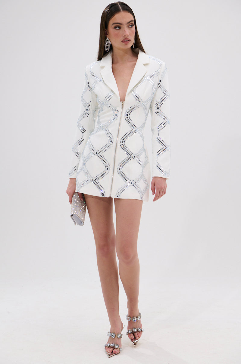 A woman wears the LOOK AT ME RHINESTONE BLAZER DRESS IN WHITE with geometric silver patterns, paired with silver earrings, embellished heels, and a small silver clutch showing its satin-lined interior against a white background.