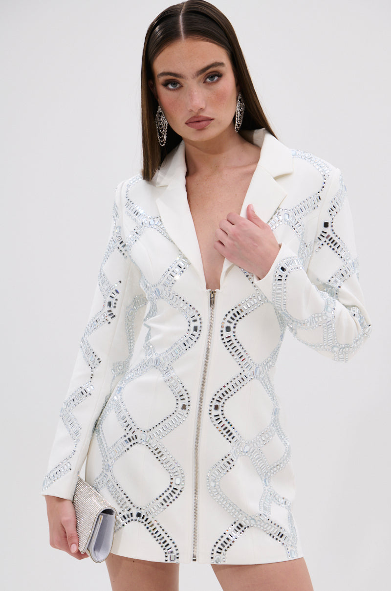 A woman wears the LOOK AT ME RHINESTONE BLAZER DRESS IN WHITE, a fitted white dress with silver embellishments and satin lining. She carries a sparkling silver clutch, her long brown hair styled with dramatic makeup and statement earrings.