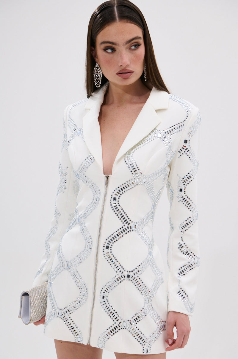 A woman in a white LOOK AT ME RHINESTONE BLAZER DRESS stands against a plain background, featuring silver geometric embellishments, a satin lining, statement earrings, and a sparkly clutch.