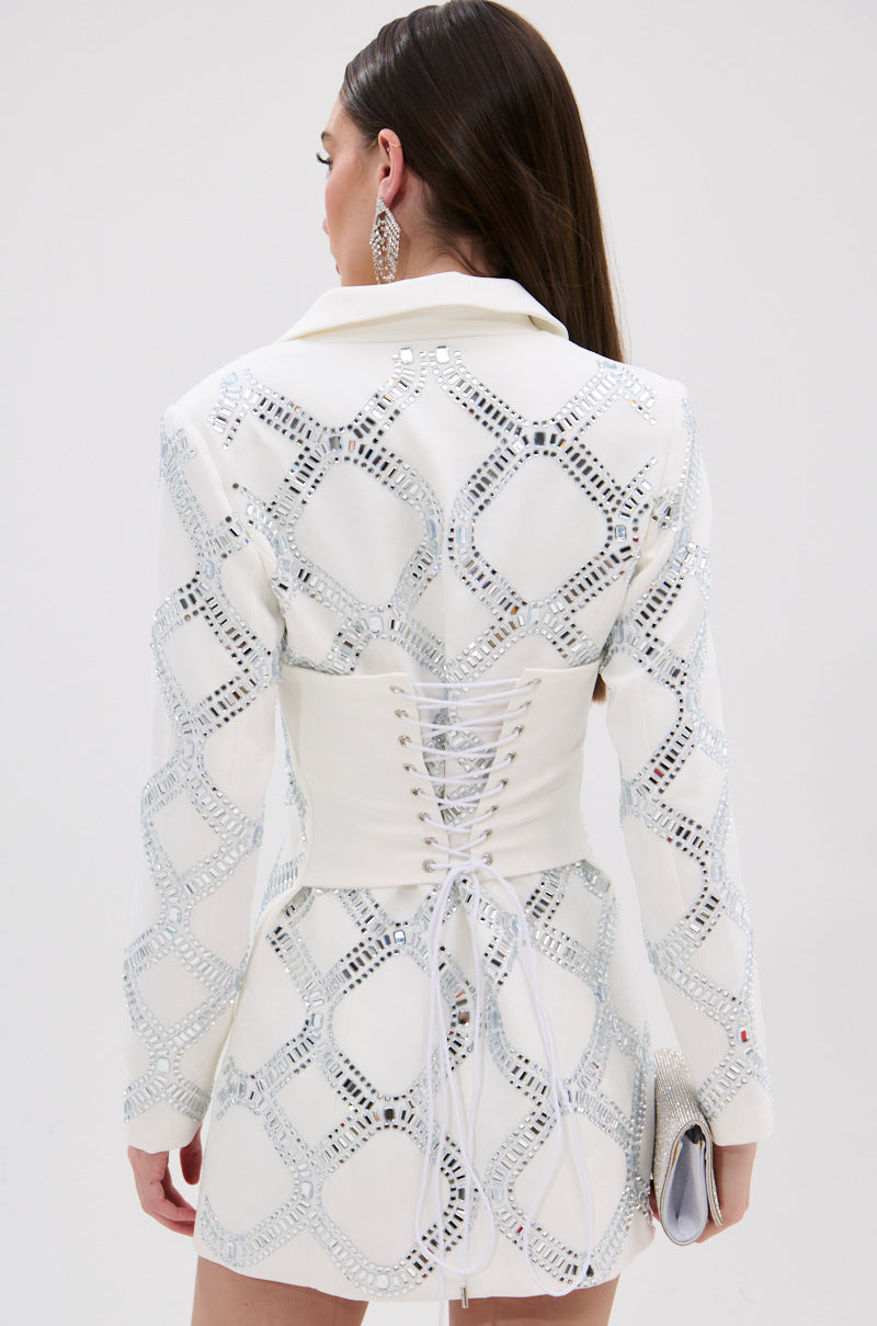 A woman with long brown hair is seen from behind wearing the LOOK AT ME RHINESTONE BLAZER DRESS IN WHITE, which features silver geometric rhinestones, a satin-lined interior, and a white lace-up corset back. She holds a clutch and wears large earrings.