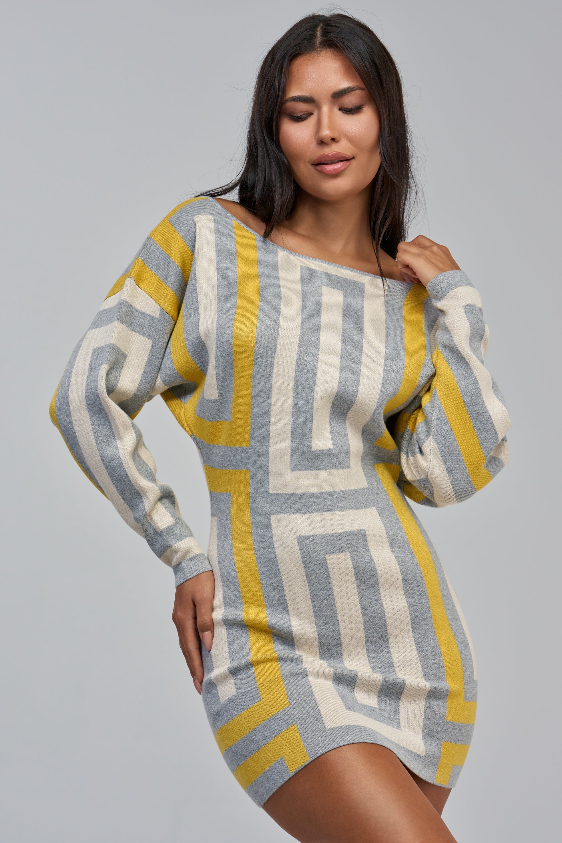 Model showcasing the PRETTY & PAID MINI SWEATER DRESS, a long-sleeve, form-fitting sweater dress featuring a bold geometric print in yellow, gray, and white. She poses against a plain gray background with one hand near her hair, creating a chic statement daytime look.