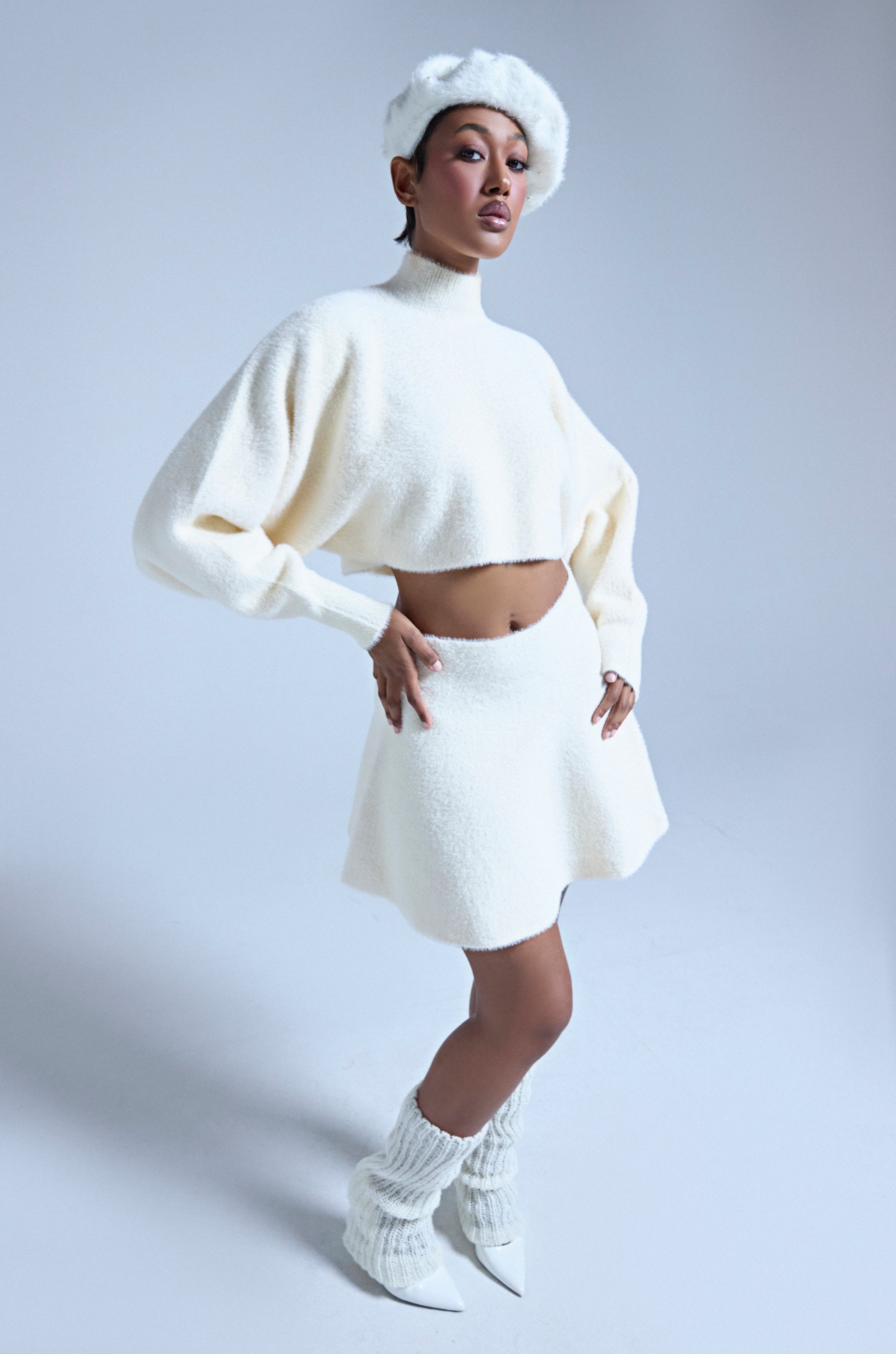 A person stands confidently against a light background wearing the CUDDLE UP FUZZY MINI SKIRT IN IVORY, styled with a cozy mohair-like cropped sweater, beret, fuzzy leg warmers, and pointed white heels. 0