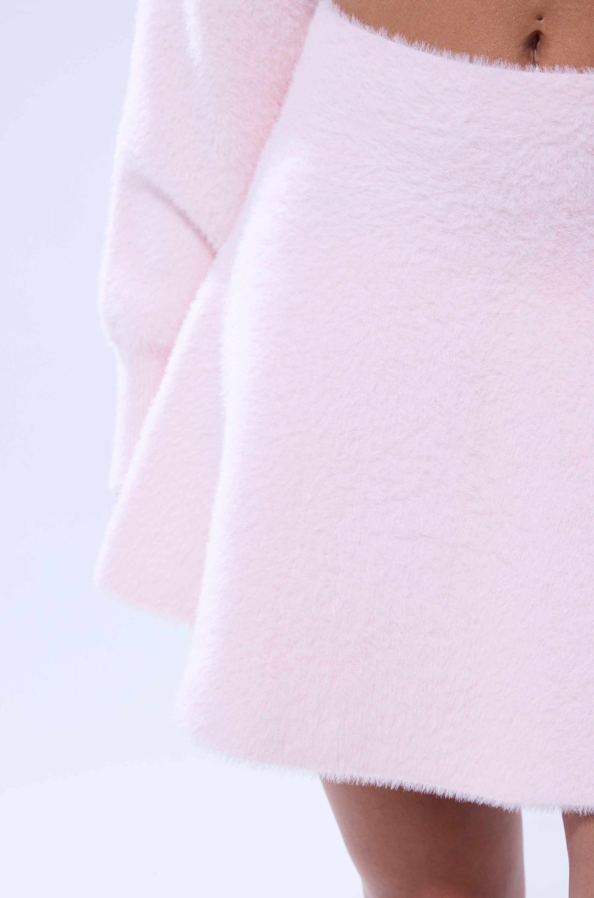 Close-up of a person wearing the CUDDLE UP FUZZY MINI SKIRT IN PINK, a soft, high-rise skirt, paired with a matching textured top. Only part of their lower body is visible against a pale background. 6