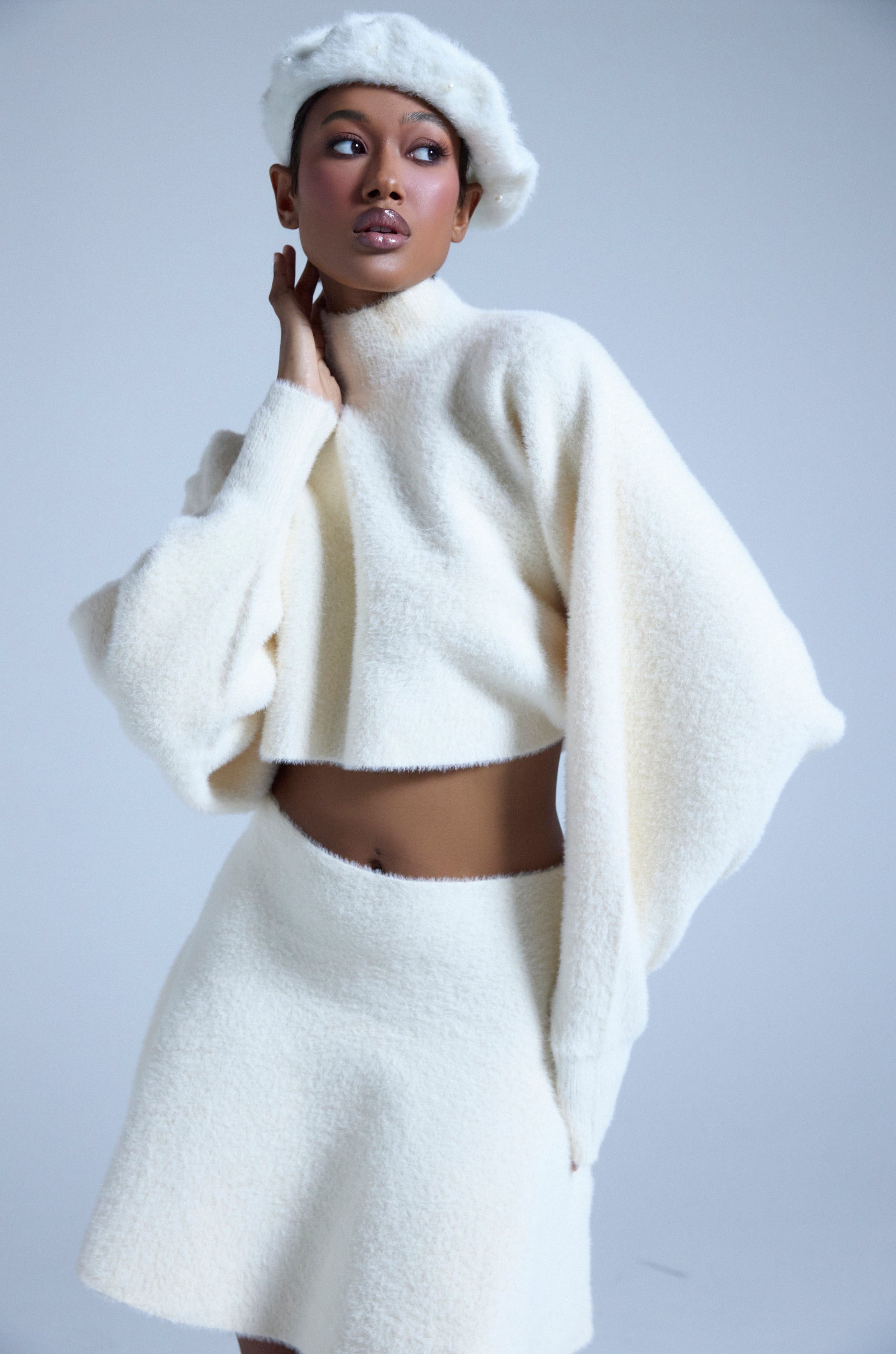 A woman models the CUDDLE UP FUZZY CROPPED SWEATER IN IVORY with a matching skirt and beret, posing against a plain light background and gazing to the side with one hand at her neck.