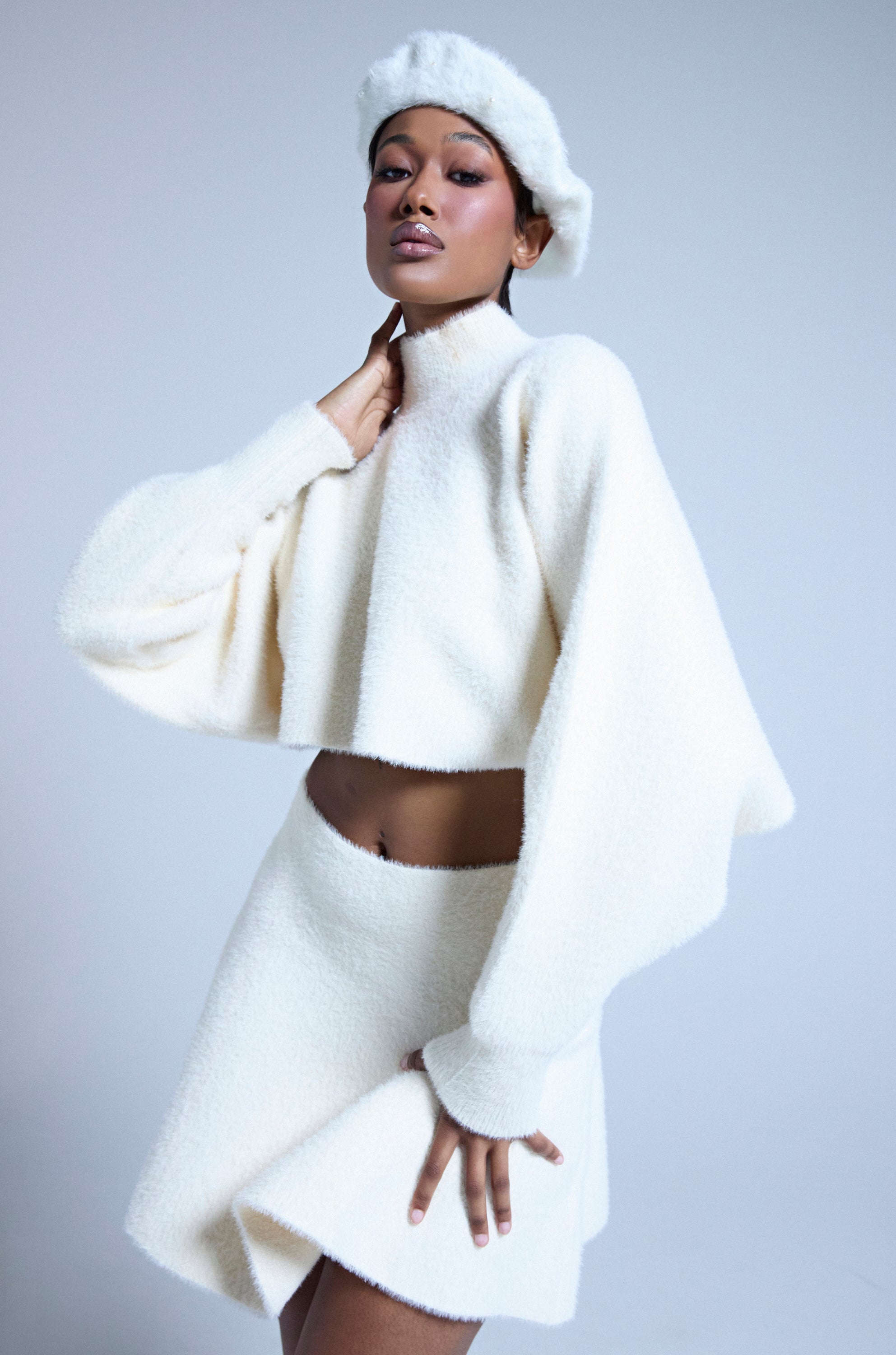 A person poses confidently in the CUDDLE UP FUZZY CROPPED SWEATER IN IVORY, styled with a matching mini skirt and beret. The mohair-like fabric and soft textures create a chic, monochrome look against a plain grey background.