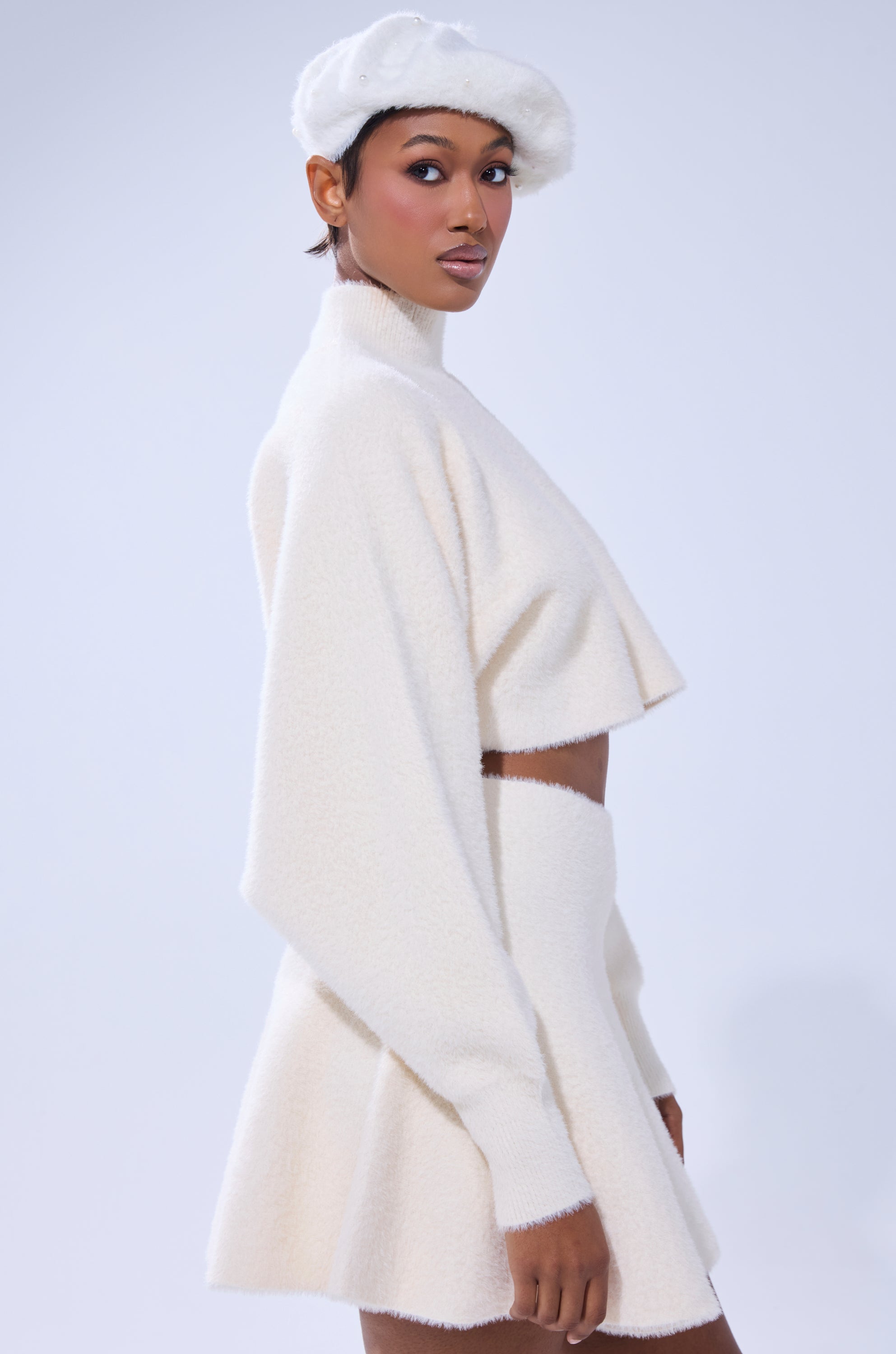 A woman poses in the CUDDLE UP FUZZY CROPPED SWEATER IN IVORY and a matching skirt, styled with a white beret, against a light background.