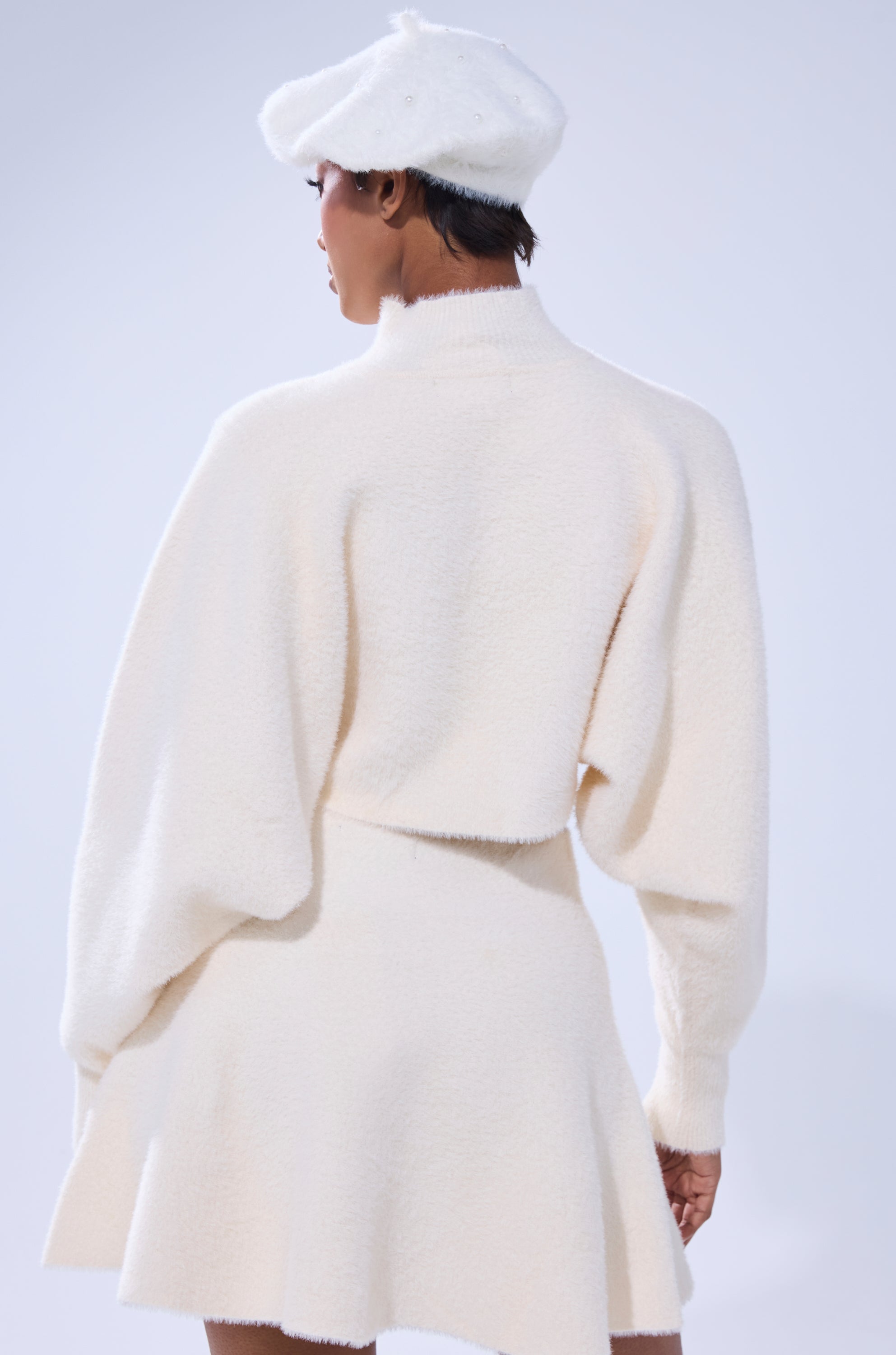 A person is seen from behind wearing the CUDDLE UP FUZZY CROPPED SWEATER IN IVORY, paired with a matching skirt and white beret. The mohair-like fabric enhances the cozy, chic feel against a plain light gray background.