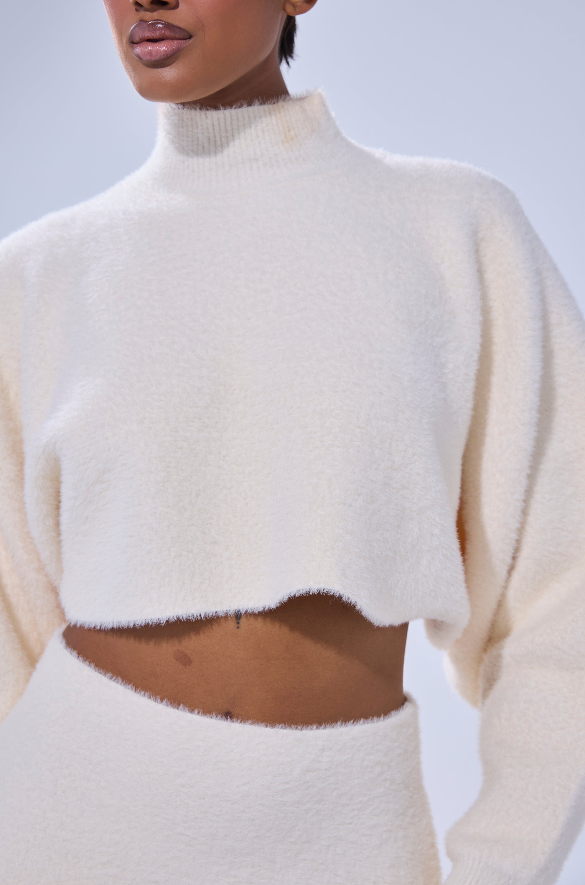 A person models the CUDDLE UP FUZZY CROPPED SWEATER IN IVORY, featuring long sleeves and a high neckline in mohair-like fabric, styled with a matching high-waisted skirt for a chic loungewear look that shows off the midriff.