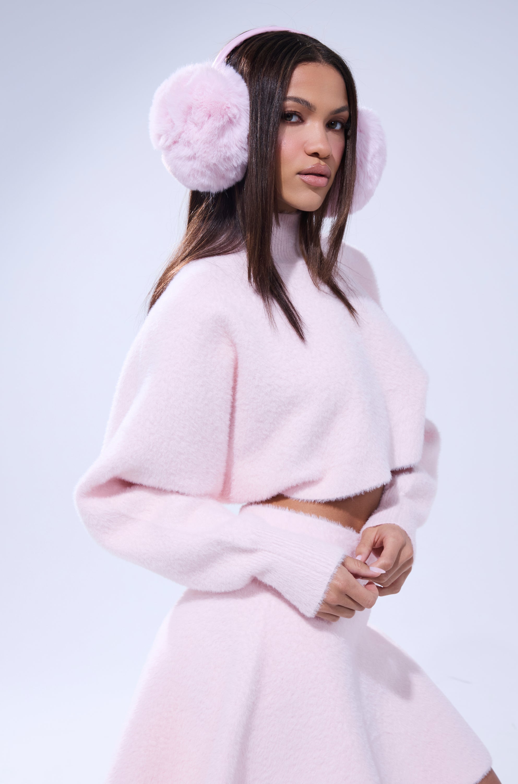 A woman with straight brown hair and a confident look wears the CUDDLE UP FUZZY CROPPED SWEATER IN PINK with a matching skirt and fluffy earmuffs, posing against a simple light background.