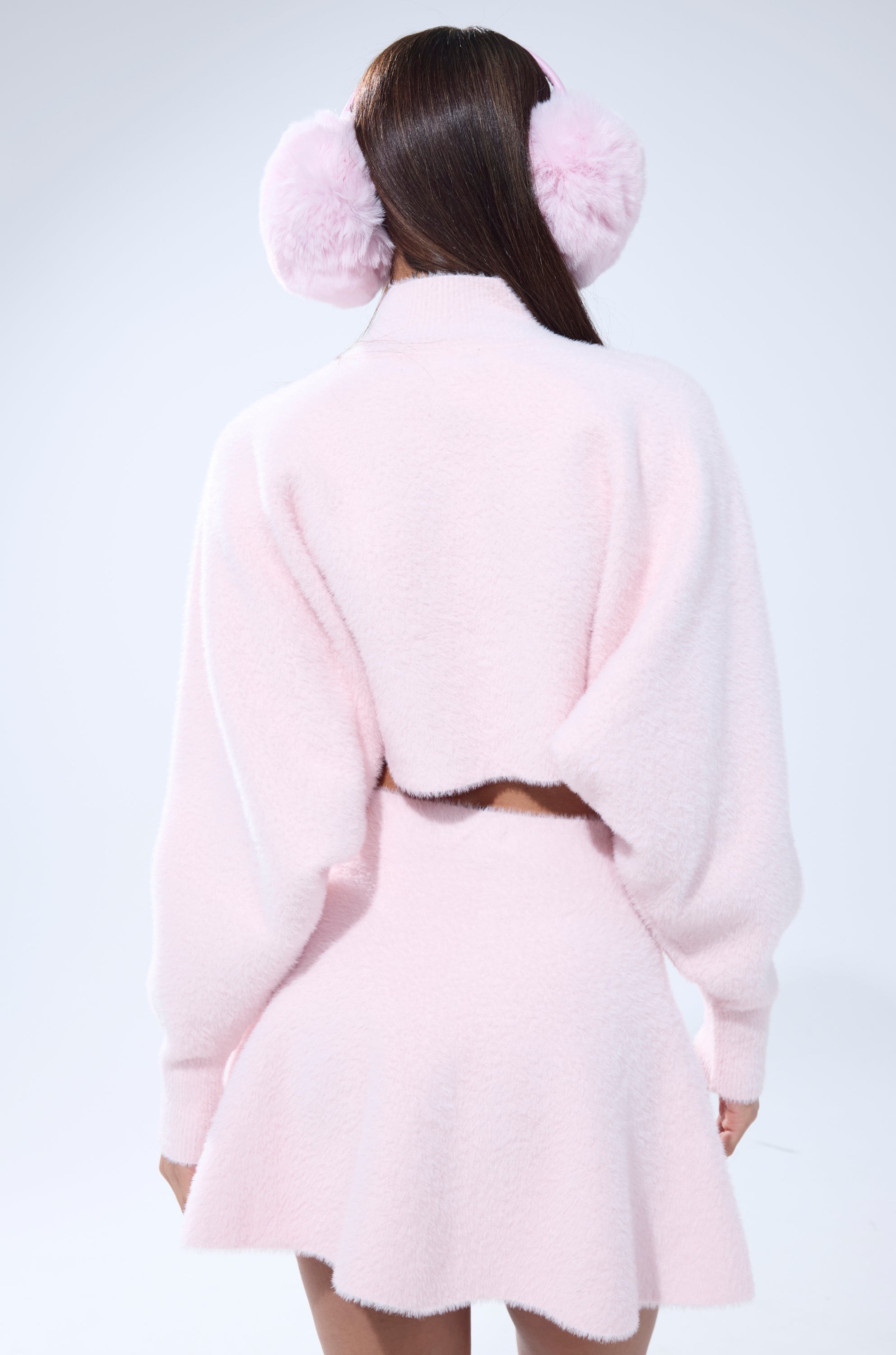 A person with long brown hair faces away from the camera against a light background, wearing the CUDDLE UP FUZZY CROPPED SWEATER IN PINK along with fluffy pink earmuffs and a matching pastel pink skirt in mohair-like fabric.