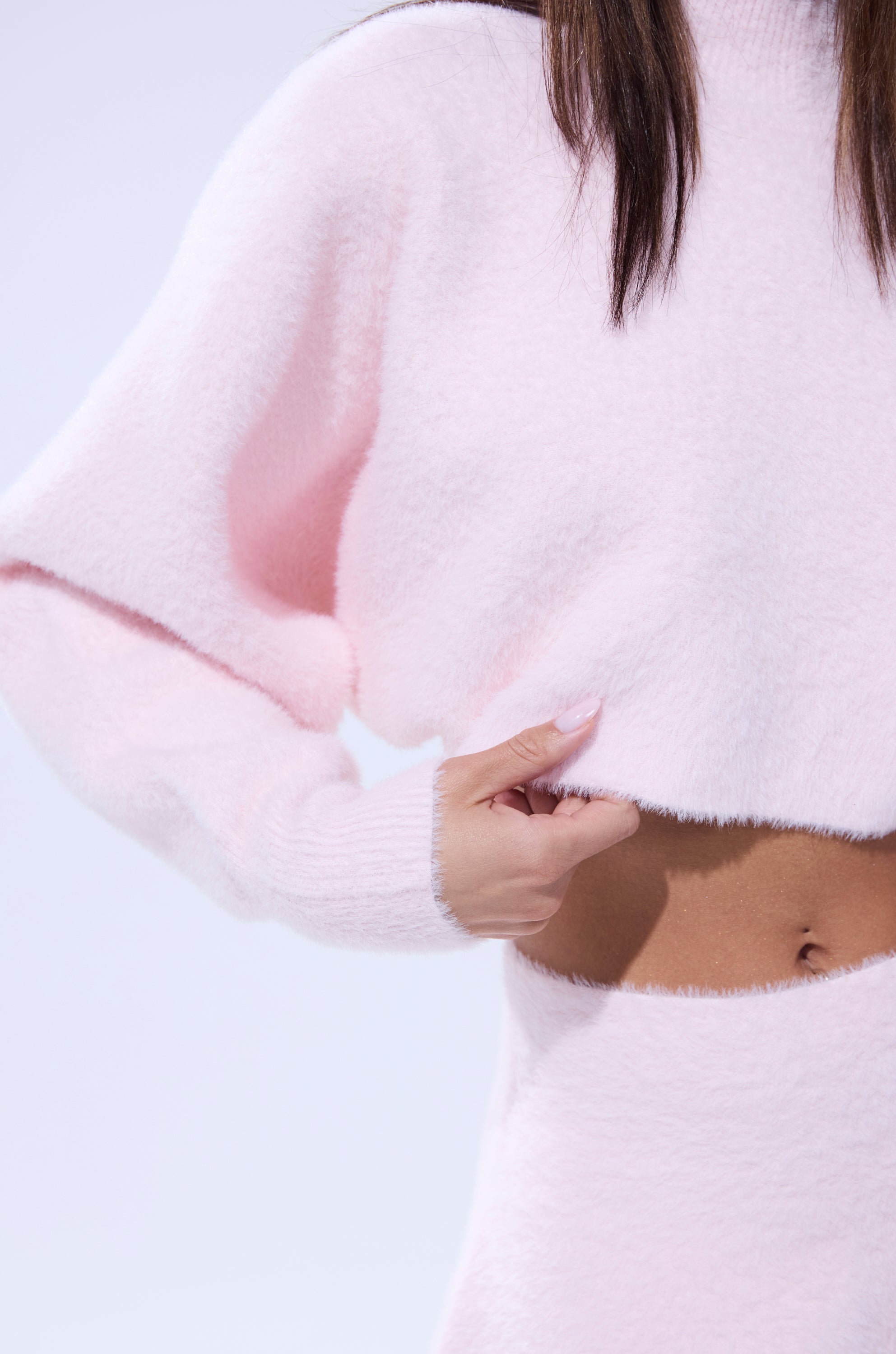 Wearing the CUDDLE UP FUZZY CROPPED SWEATER IN PINK, a person gently lifts the soft mohair-like top to reveal their midriff, its light pink hue standing out against a plain background.