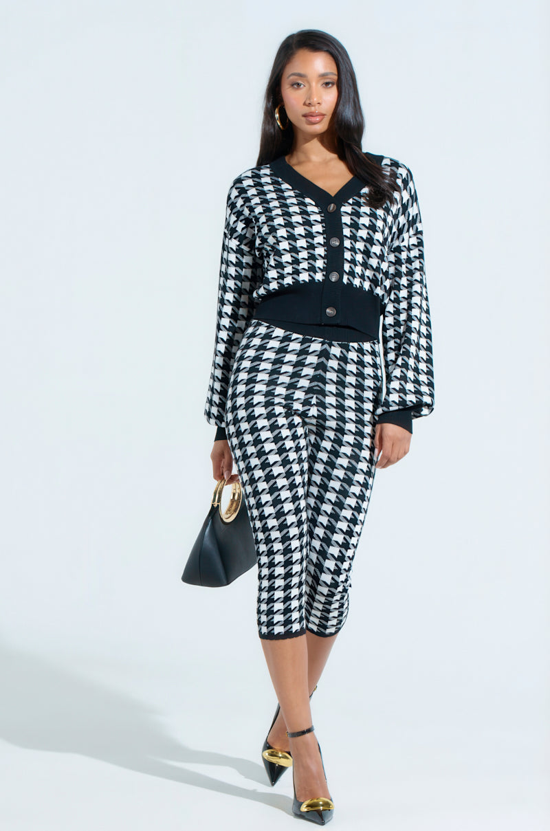 A woman exudes confidence in a HIGH SOCIETY CAPRI houndstooth cardigan and skirt outfit made from stretchy knit. She accessorizes with a black handbag and heels, set against a plain light backdrop.