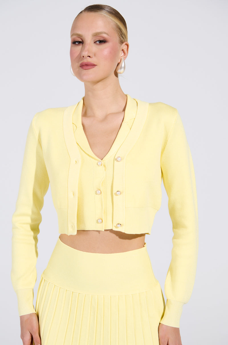 A person poses against a plain backdrop, wearing the LEMONY EMILY Cropped Light Weight Cardigan in pastel yellow with gold-wreathed pearl buttons, over a matching top and pleated skirt. Their hair is styled neatly back.