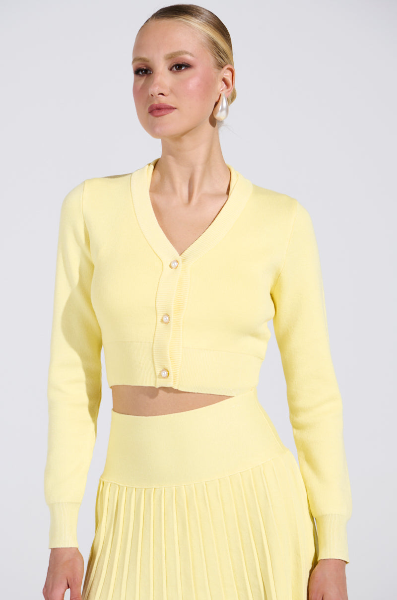 Dressed in the LEMONY EMILY cropped lightweight yellow cardigan, adorned with gold-wreathed pearl buttons, and a matching pleated skirt. The V-neckline enhances elegance, while their sleek pulled-back hair complements the plain white backdrop.