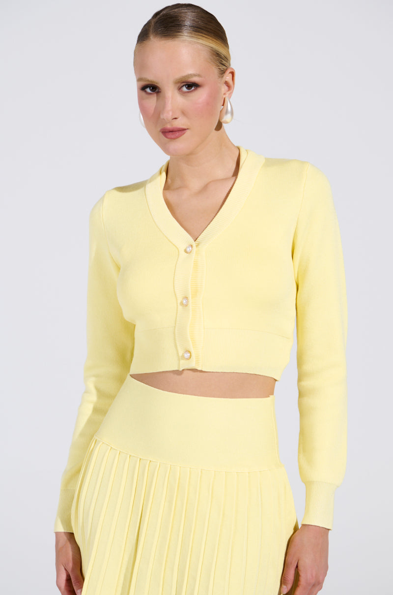 A person with slicked-back hair wears the LEMONY EMILY Cropped Lightweight Cardigan in Yellow, featuring gold-wreathed pearl buttons, paired with a matching pleated skirt. The plain background enhances the outfit's soft color.