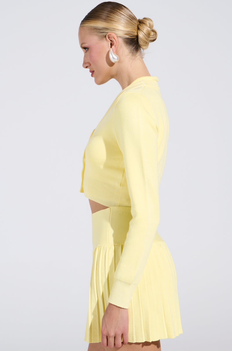 A person with blonde hair in a bun, wearing large white hoop earrings, poses sideways against a plain background in the LEMONY EMILY CROPPED LIGHT WEIGHT CARDIGAN IN YELLOW, featuring gold-wreathed pearl buttons and paired with a matching pleated skirt.