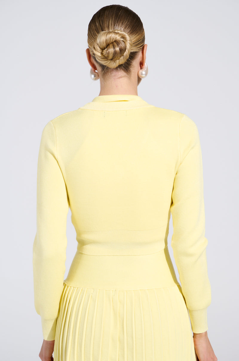 A person with blonde hair in a bun and pearl earrings wears the LEMONY EMILY cropped light weight cardigan, featuring gold-wreathed pearl buttons, over a pleated skirt. The background is plain white.