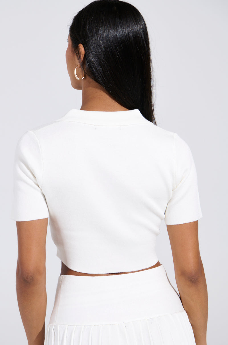 The image shows a person with long black hair from behind, wearing the Lemony Emily Button Down Top in white, a stretchy knit fabrication short-sleeved crop top and skirt, along with a small gold hoop earring on one ear.