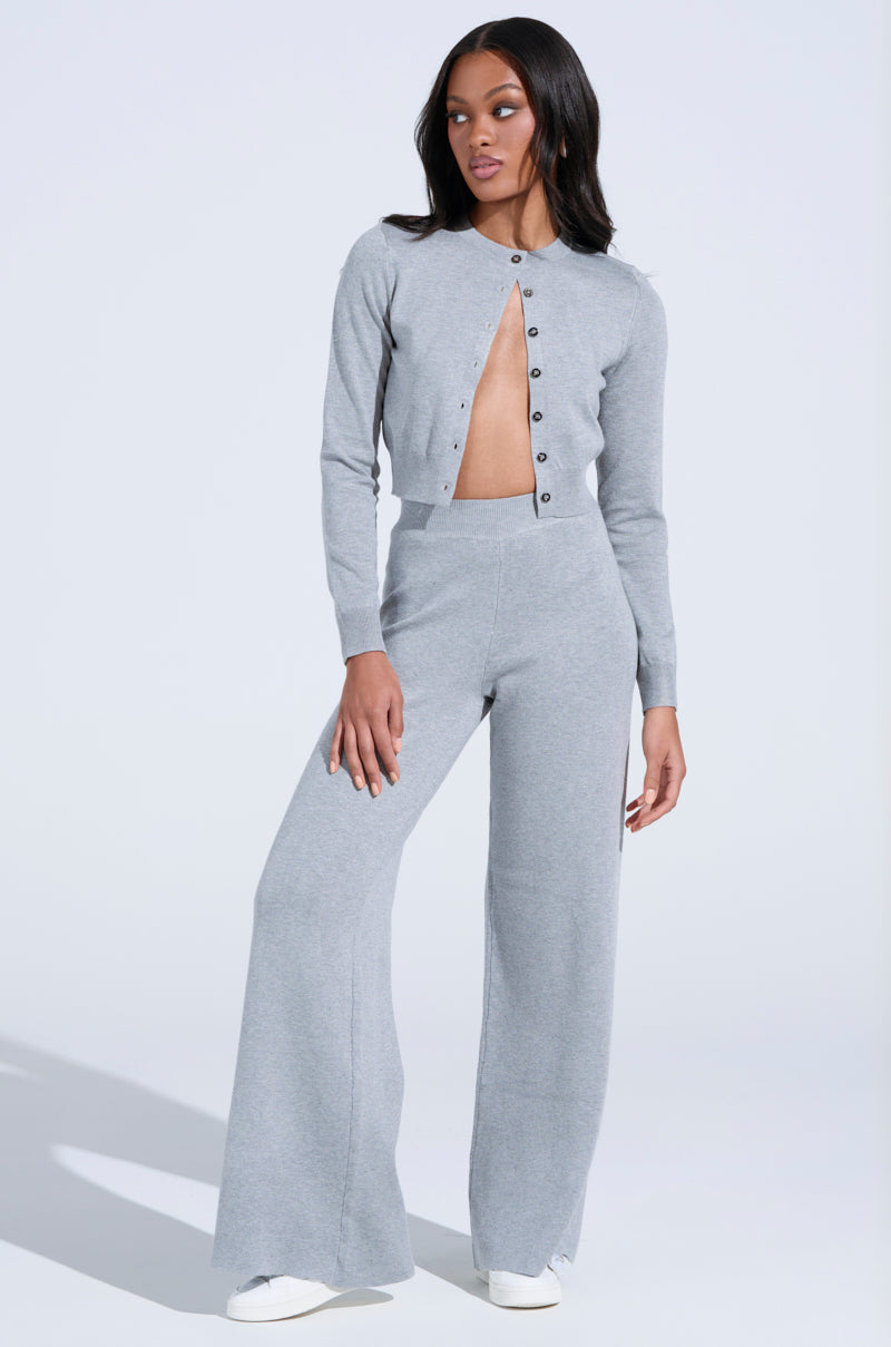 Dressed in a light gray COZY LUXE WIDE LEG PANT and matching cropped top with buttons, the person poses against a neutral backdrop. White shoes complement the cozy yet luxurious ensemble, featuring long sleeves and high-rise, wide-leg pants.