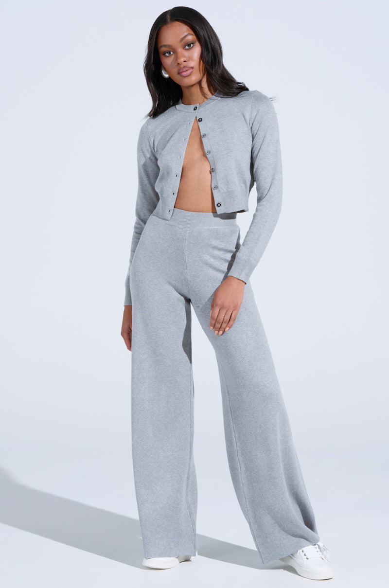 A person stands confidently wearing a light gray button-up cardigan with matching COZY LUXE WIDE LEG PANT and white sneakers. The plain background ensures the stylish, cozy ensemble is the focal point.