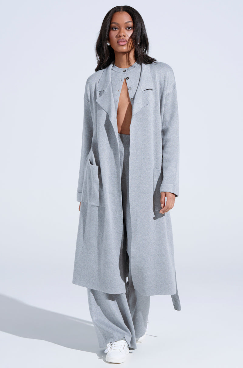 A woman walks towards the camera in a COZY LUXE KNIT TRENCH with an oversized silhouette. Her long dark hair flows over a matching low-cut top and wide-leg pants, paired with white sneakers. The self-tie belt of her coat adds elegance against the plain white background.
