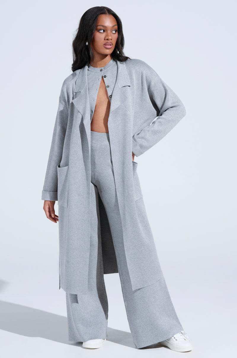 A person confidently wears a COZY LUXE KNIT TRENCH with an oversized silhouette draped over wide-leg pants and a partially buttoned top, complemented by white sneakers. The plain background accentuates the minimalist ensemble.