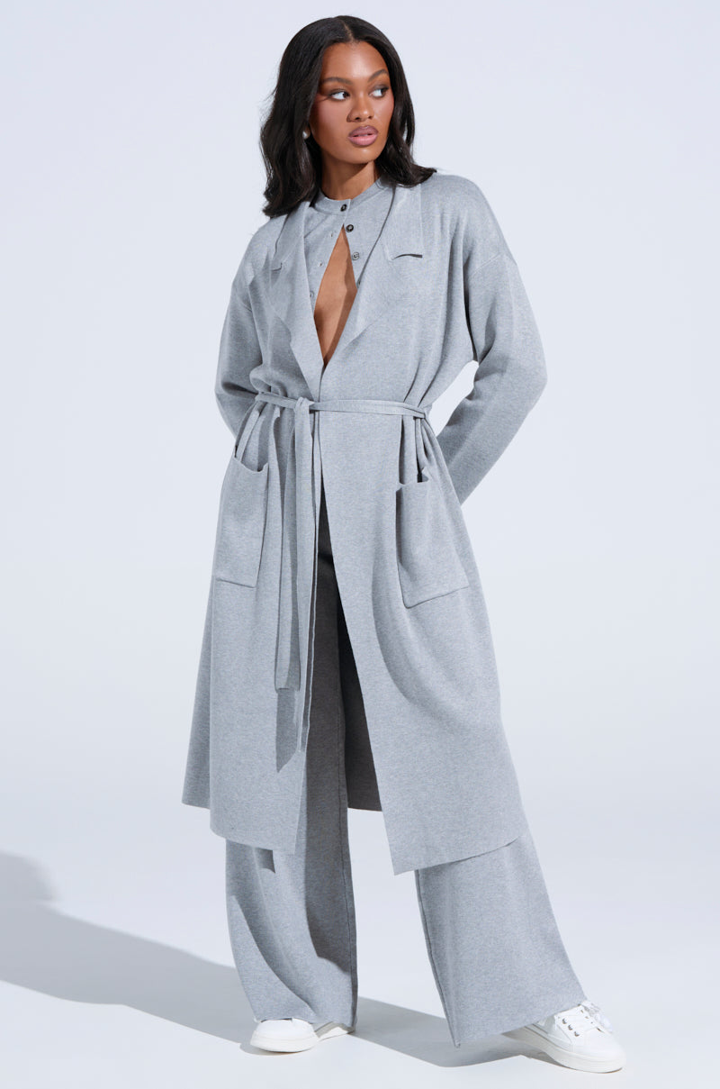 A person models an oversized silhouette with a light gray COZY LUXE KNIT TRENCH, wide open to reveal the chest, paired with matching wide-leg pants and white sneakers, set against a plain, well-lit background.
