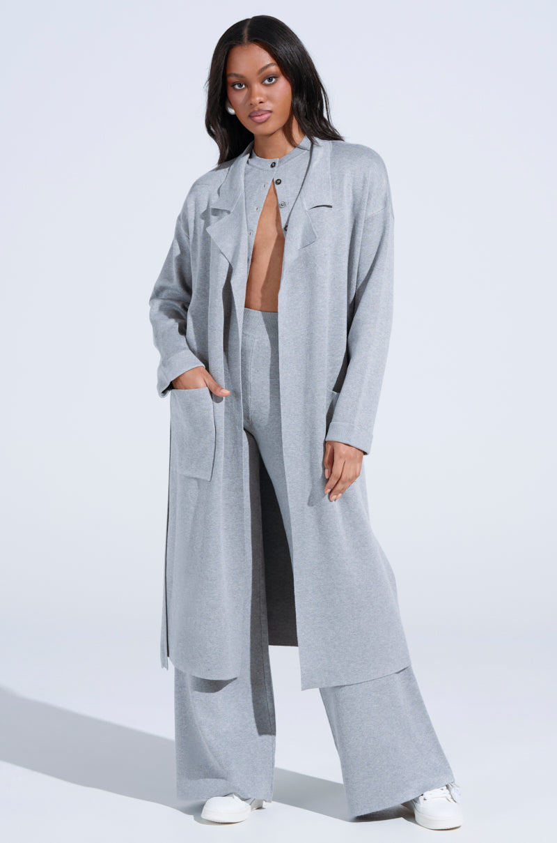 A person confidently poses in an oversized silhouette wearing a COZY LUXE KNIT TRENCH over matching wide-leg pants. The trench is buttoned at the neck, showing a bare chest, with white sneakers finishing the look. One hand rests in a pocket against a plain, light gray background.