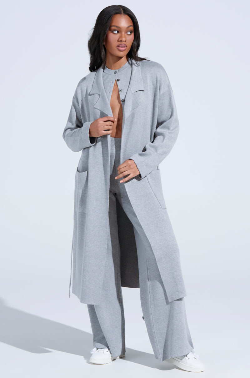 In an oversized silhouette, a person with long dark hair wears the COZY LUXE KNIT TRENCH in light gray, paired with matching pants and a partially unbuttoned shirt. They stand against a plain background, finishing the outfit with white sneakers.