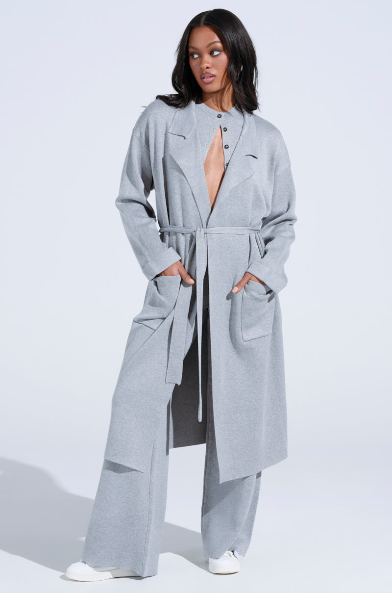 Sporting the COZY LUXE KNIT TRENCH, someone poses confidently against a plain backdrop. The oversized silhouette with a self-tie belt drapes over wide-leg pants and white sneakers, showcasing its deep V-neck and buttons.