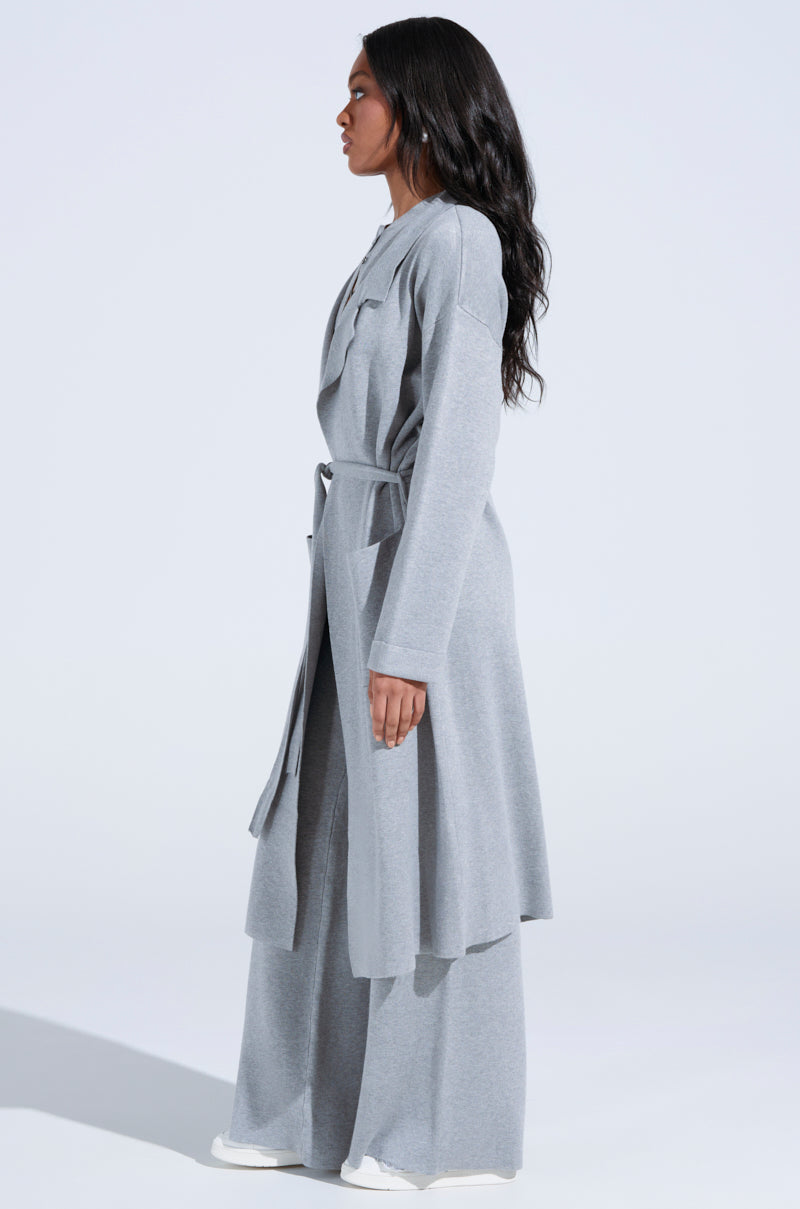 A person with long black hair stands in profile, highlighting the oversized silhouette of the COZY LUXE KNIT TRENCH in light gray, cinched with a self-tie belt over matching wide-leg pants and white sneakers. The minimalist background enhances the ensemble's elegance.